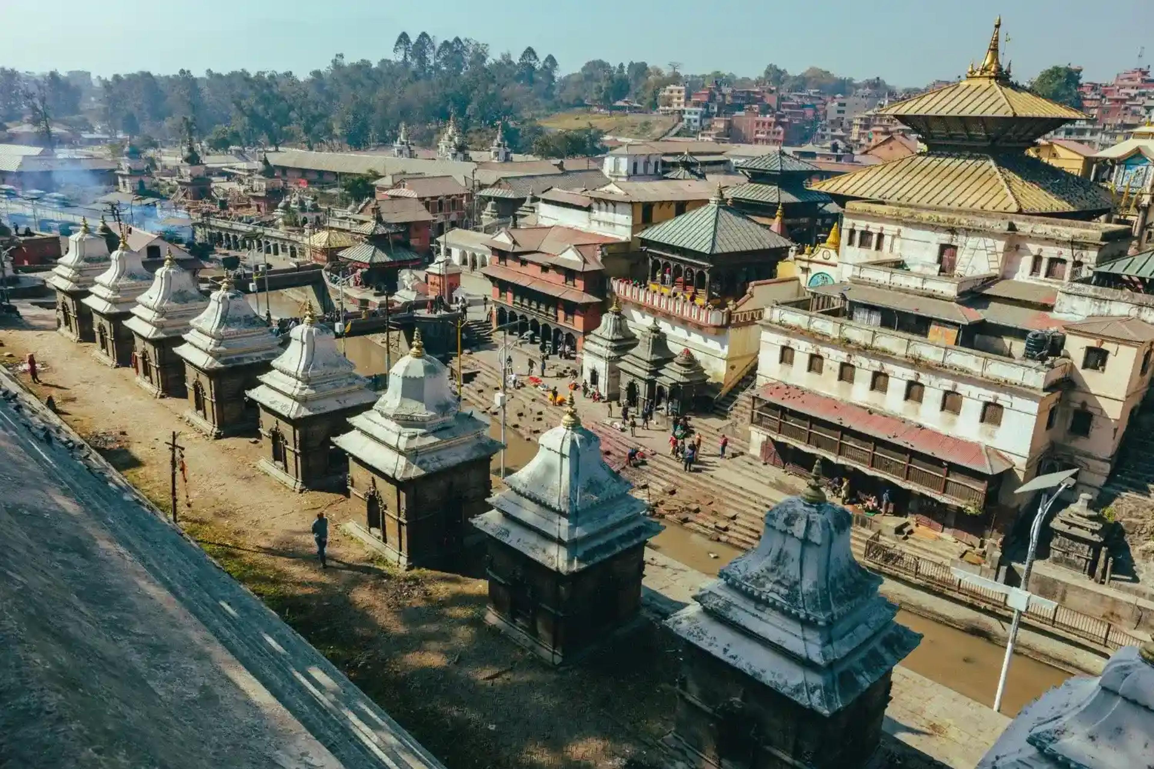 Panoramic view of the sacred Pashupatinath Temple complex in Kathmandu, showcasing white stone Shiva shrines along the Bagmati River and traditional golden-roofed pagodas.