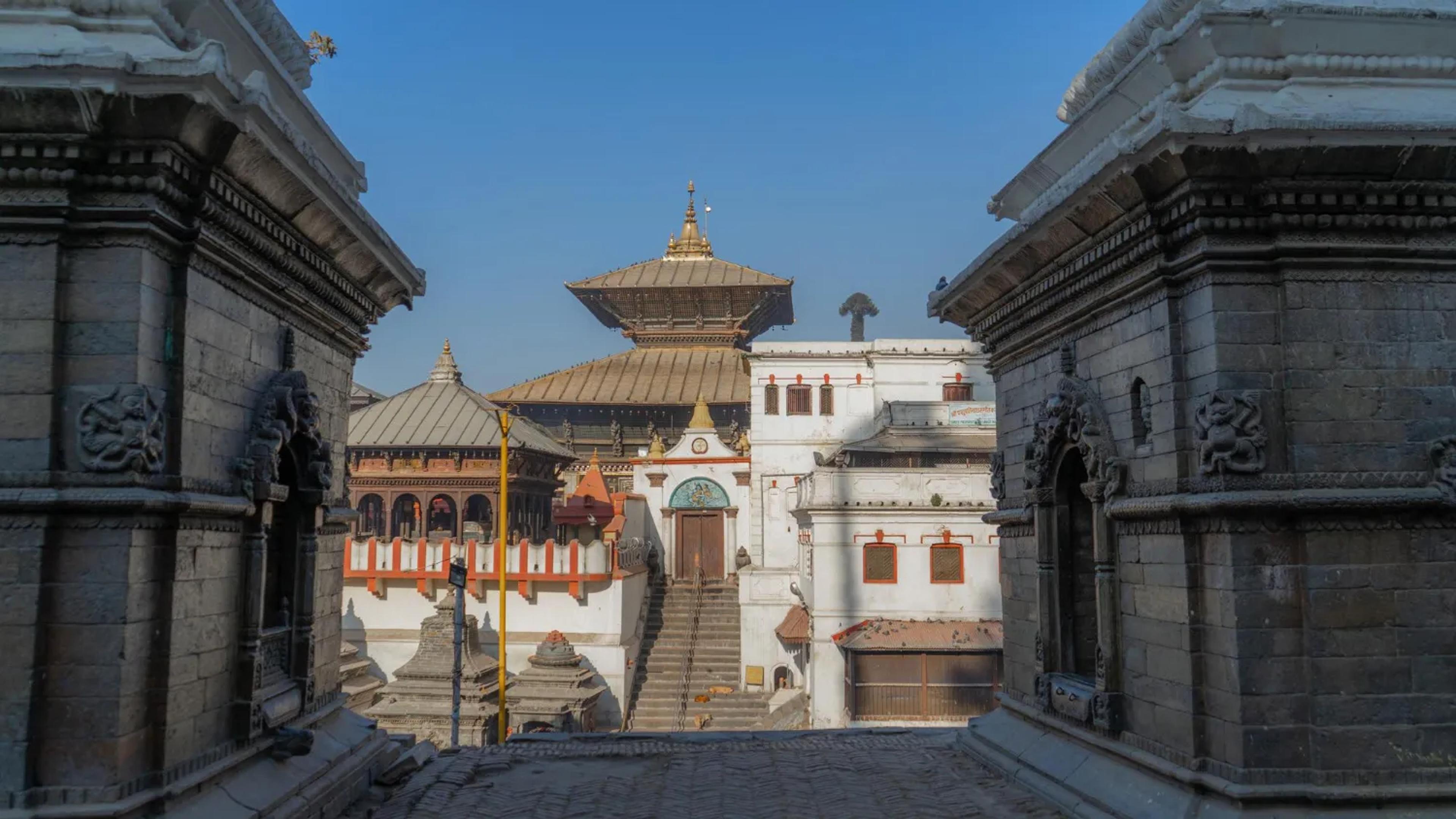 The golden tiered roof of the main Pashupatinath Temple in Kathmandu, perfectly framed between two intricately carved, ancient stone structures under a clear blue sky.
