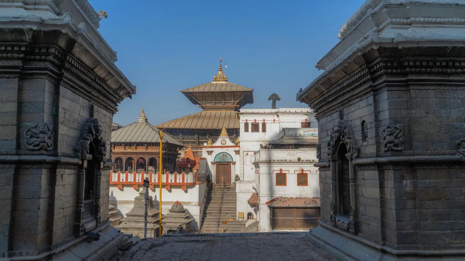 The golden tiered roof of the main Pashupatinath Temple in Kathmandu, perfectly framed between two intricately carved, ancient stone structures under a clear blue sky.