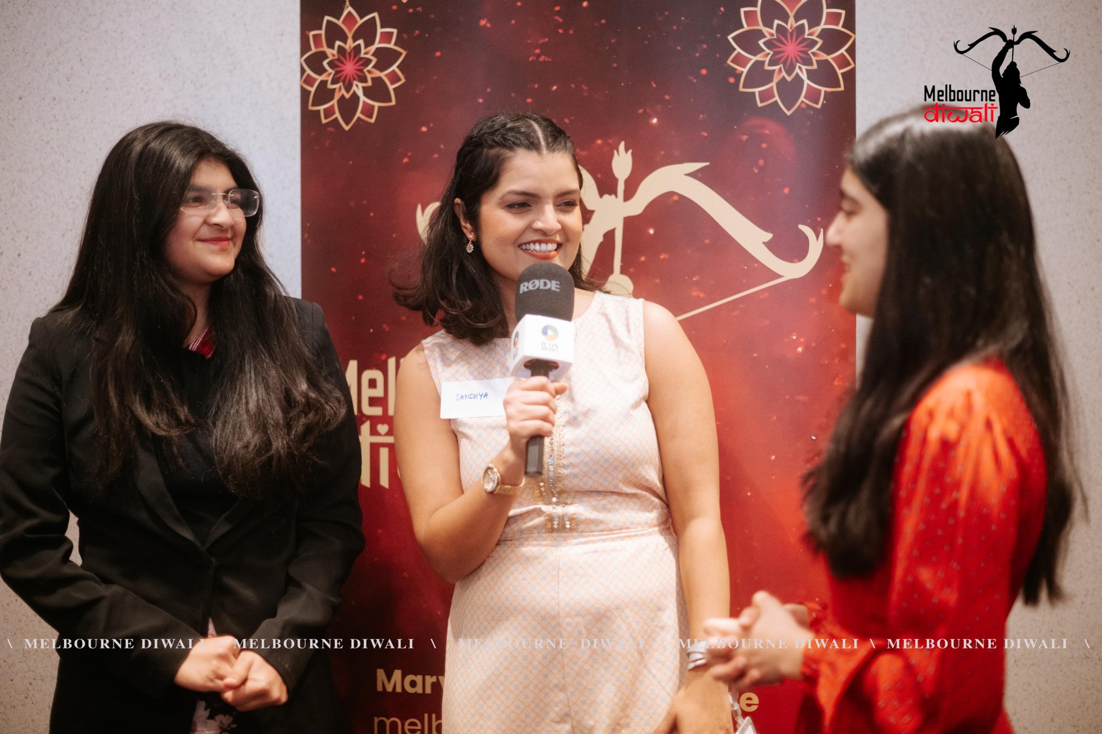 Three guests talk in front of the Melbourne Diwali media wall, one holding a microphone