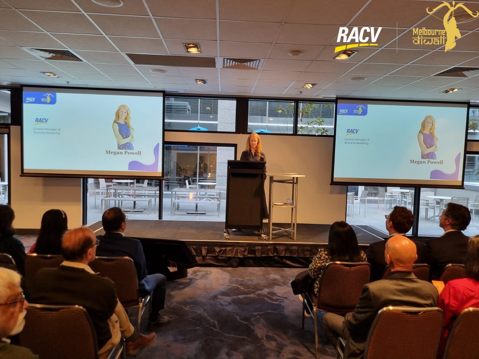 Megan Powell of RACV at the lectern, with her name slide on the twin screens