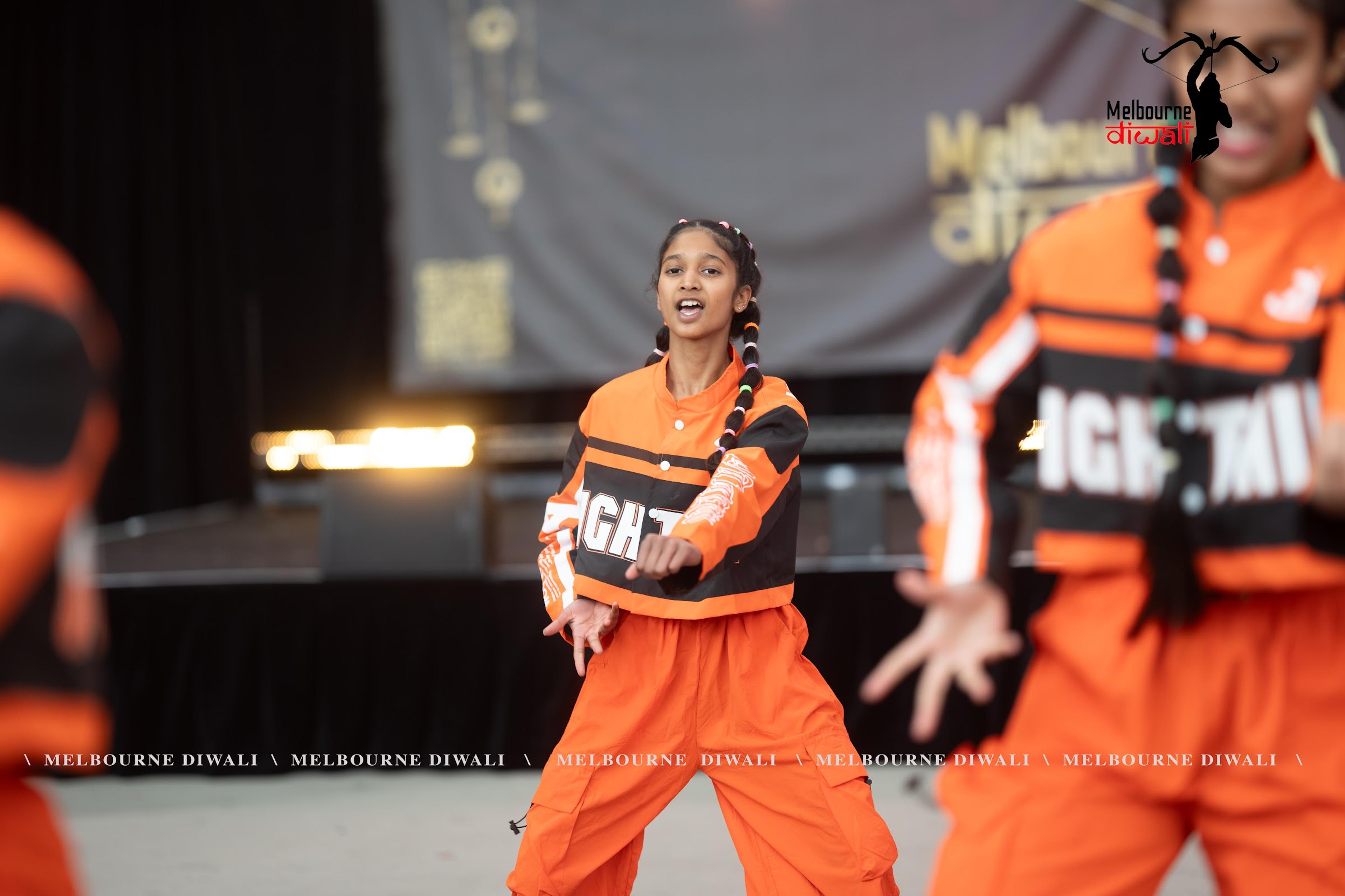 A dance crew in orange streetwear mid-routine on the main stage at Melbourne Diwali 2025