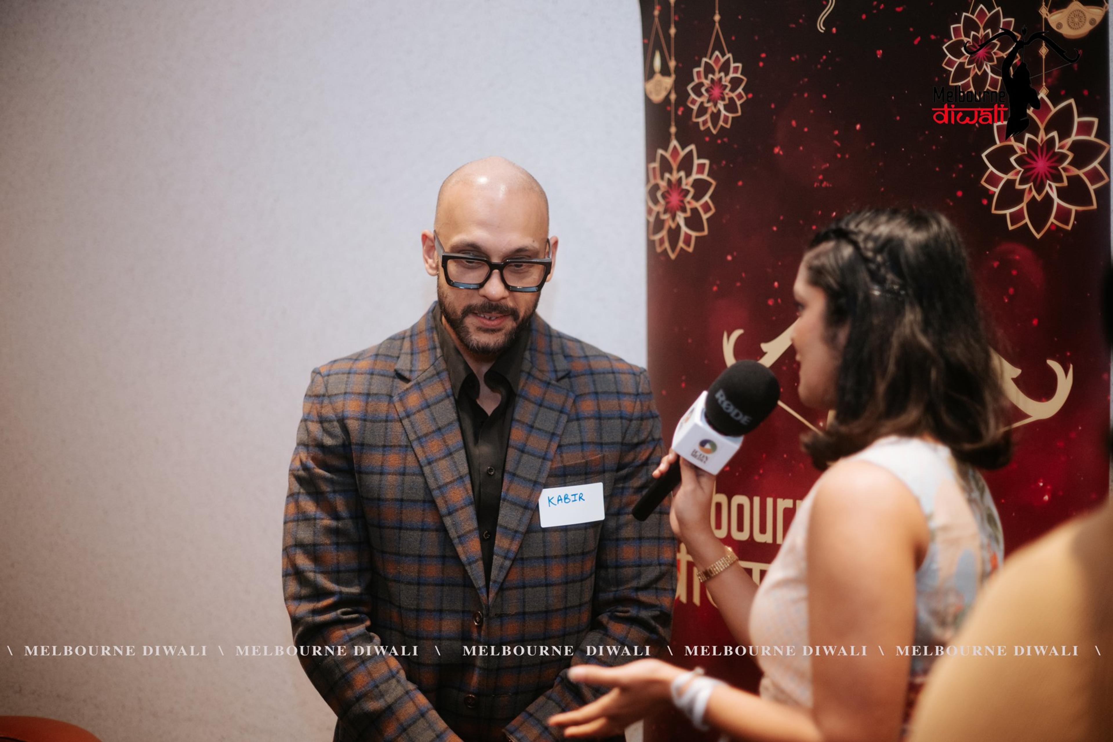 A guest in a checked blazer is interviewed with a handheld microphone in front of the Melbourne Diwali media wall