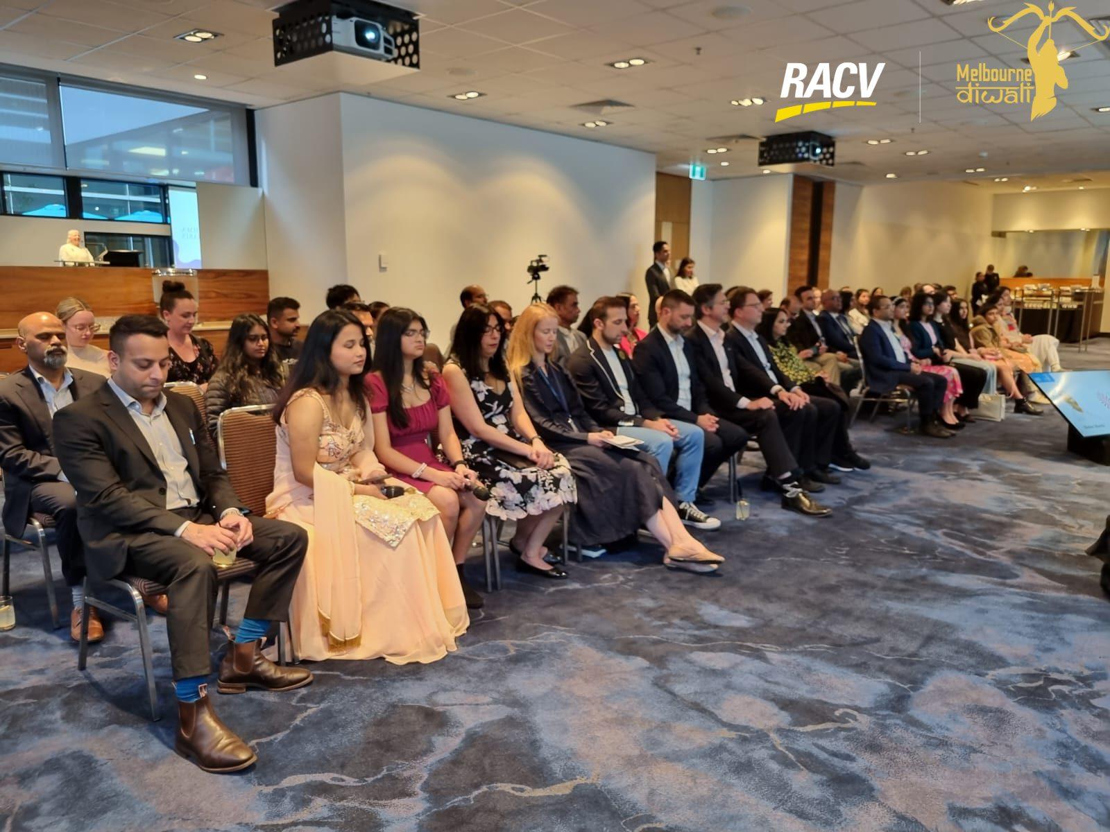 Guests seated in rows facing the stage, with a projector and a camera on a tripod in the aisle