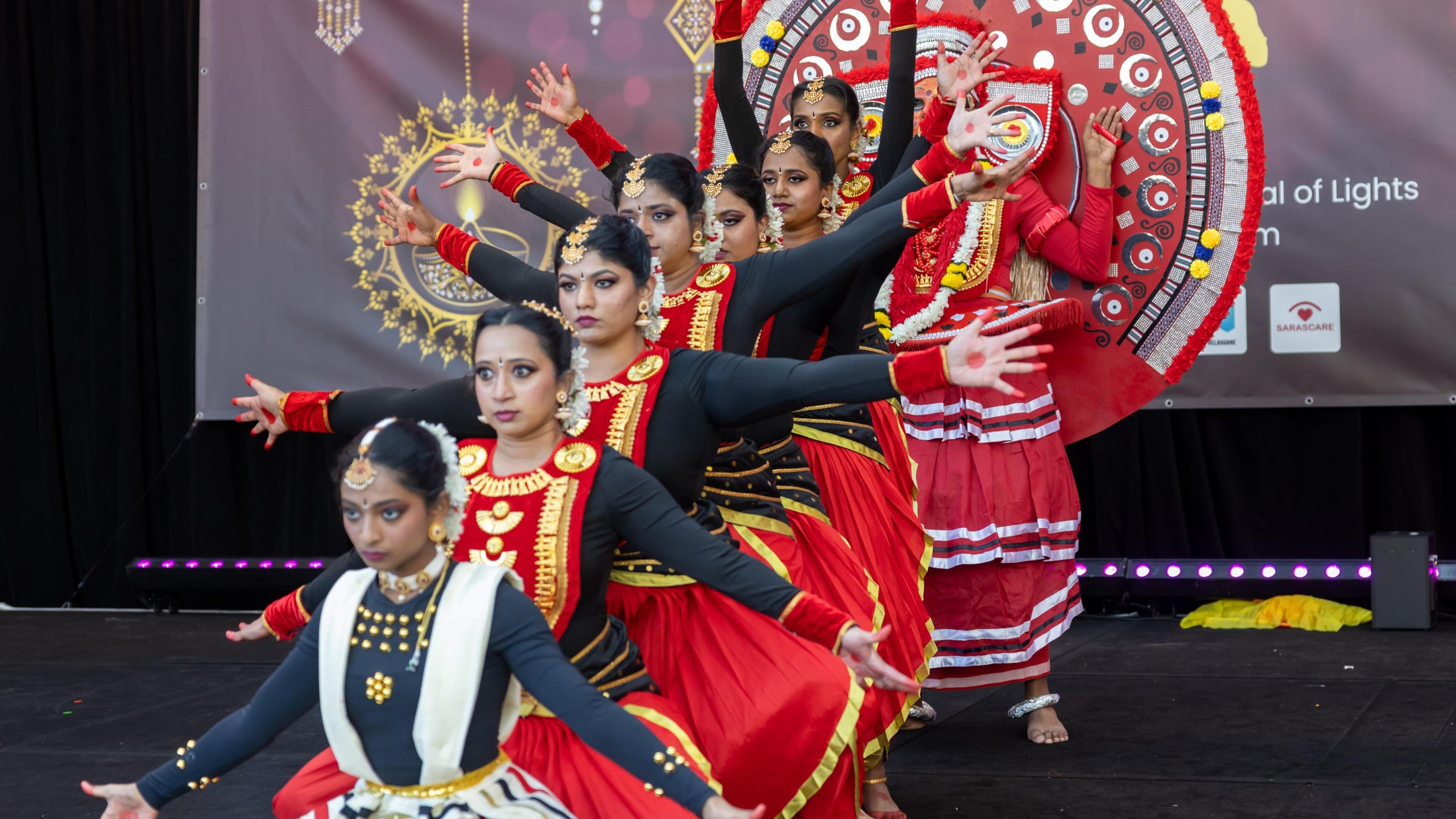 Dancers in red and black costumes performing in formation on the Melbourne Diwali stage, with a Theyyam performer behind them