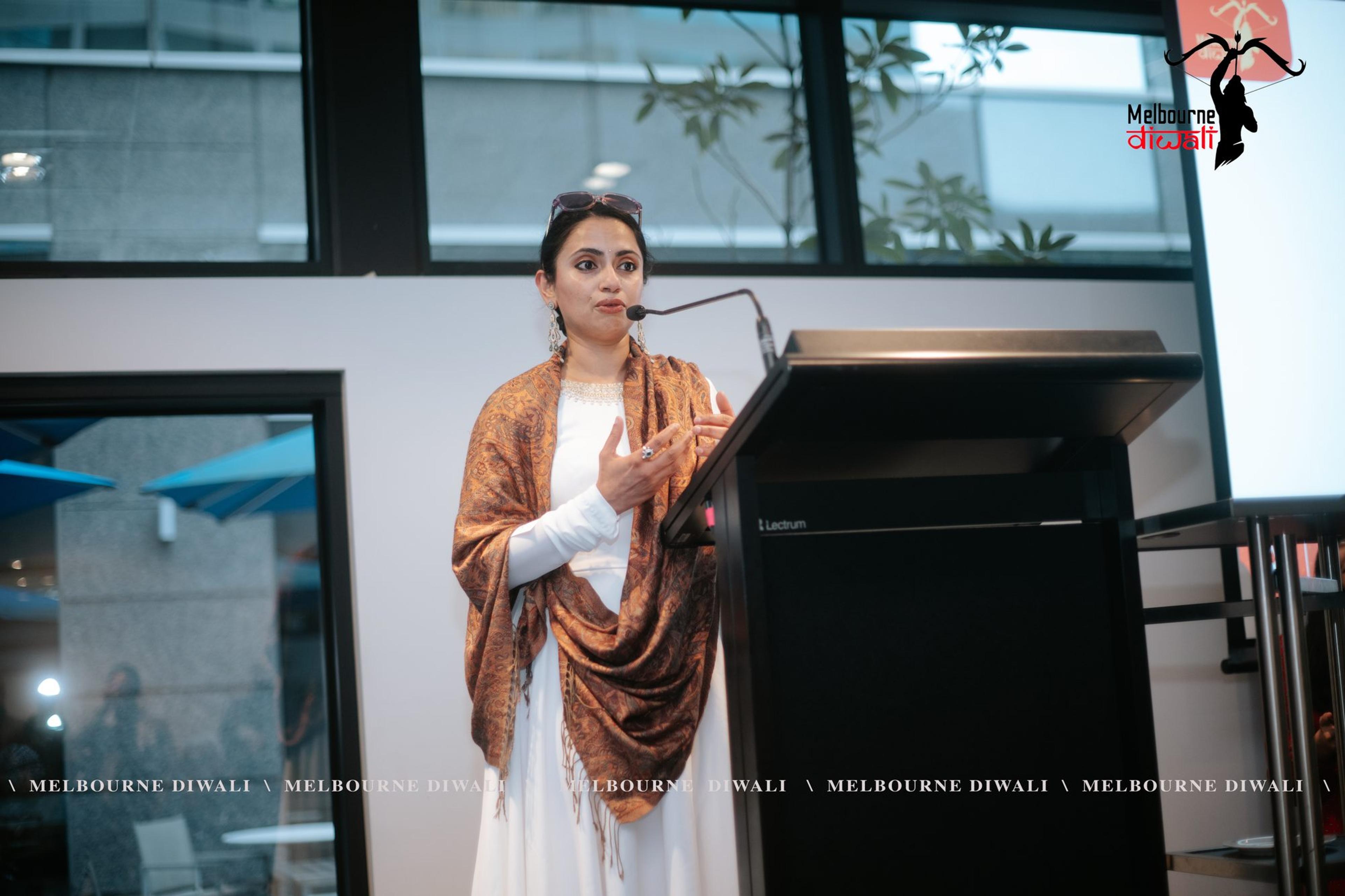 A speaker in white with a patterned shawl addresses guests at the lectern during the partner showcase