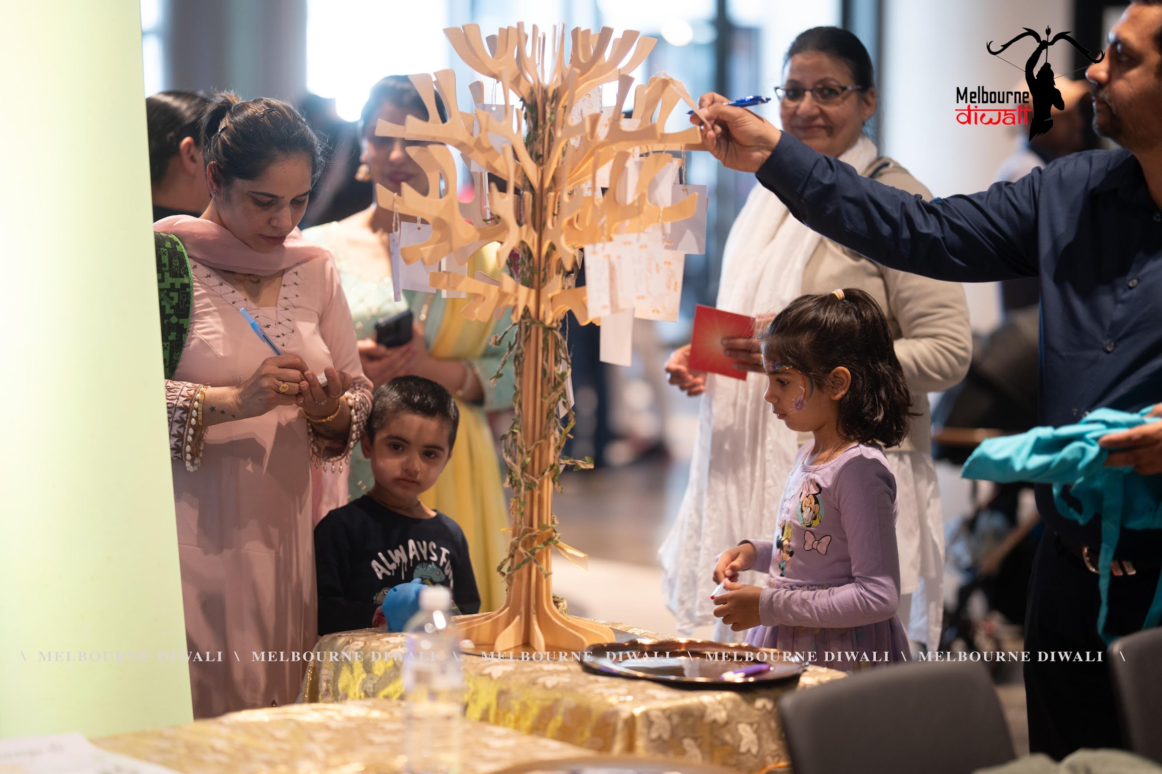 A visitor reaches up to tie a wish to the golden blessing tree while a child watches, Melbourne Diwali 2025