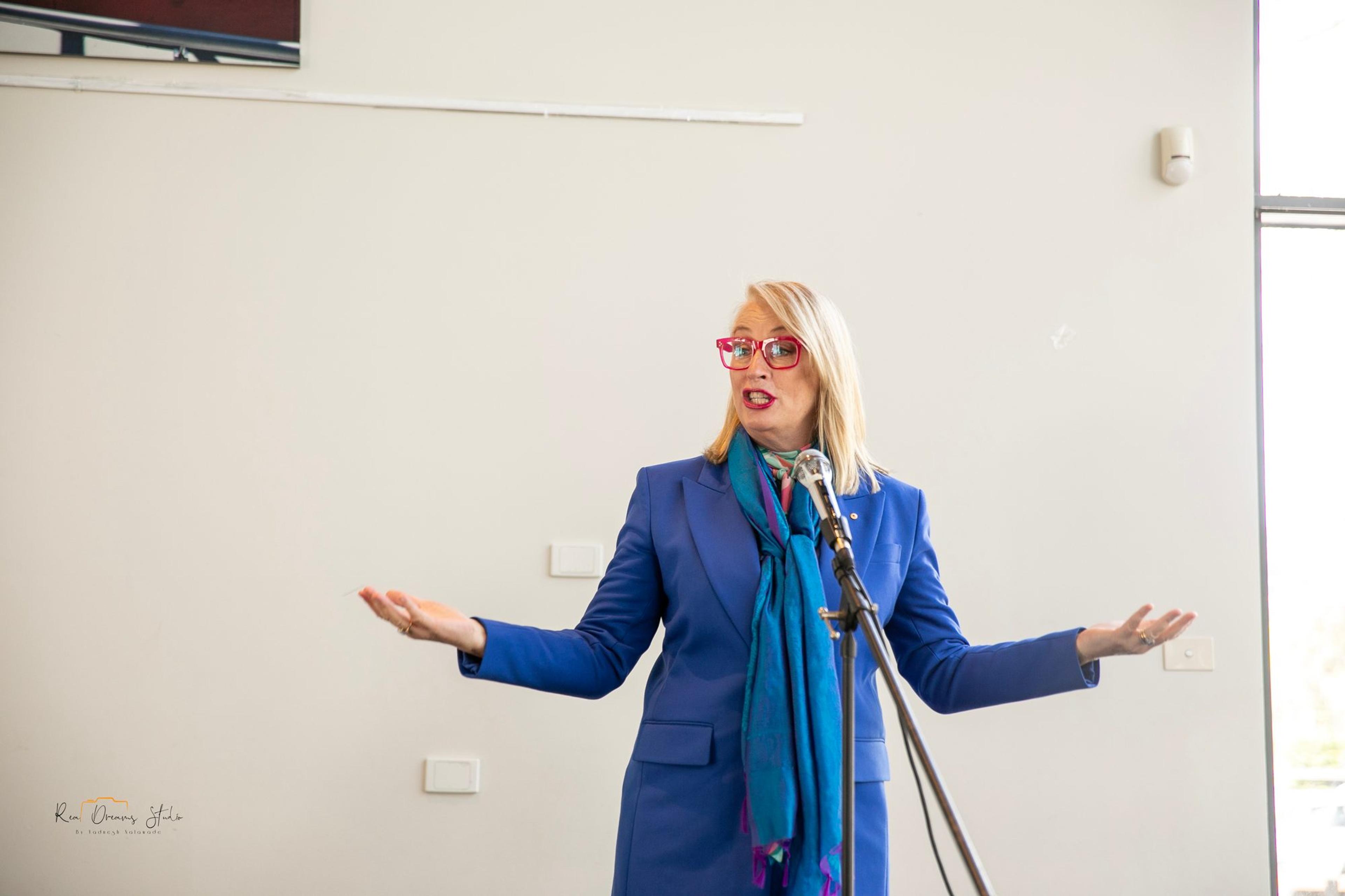 A guest in a blue jacket addresses the room, arms open