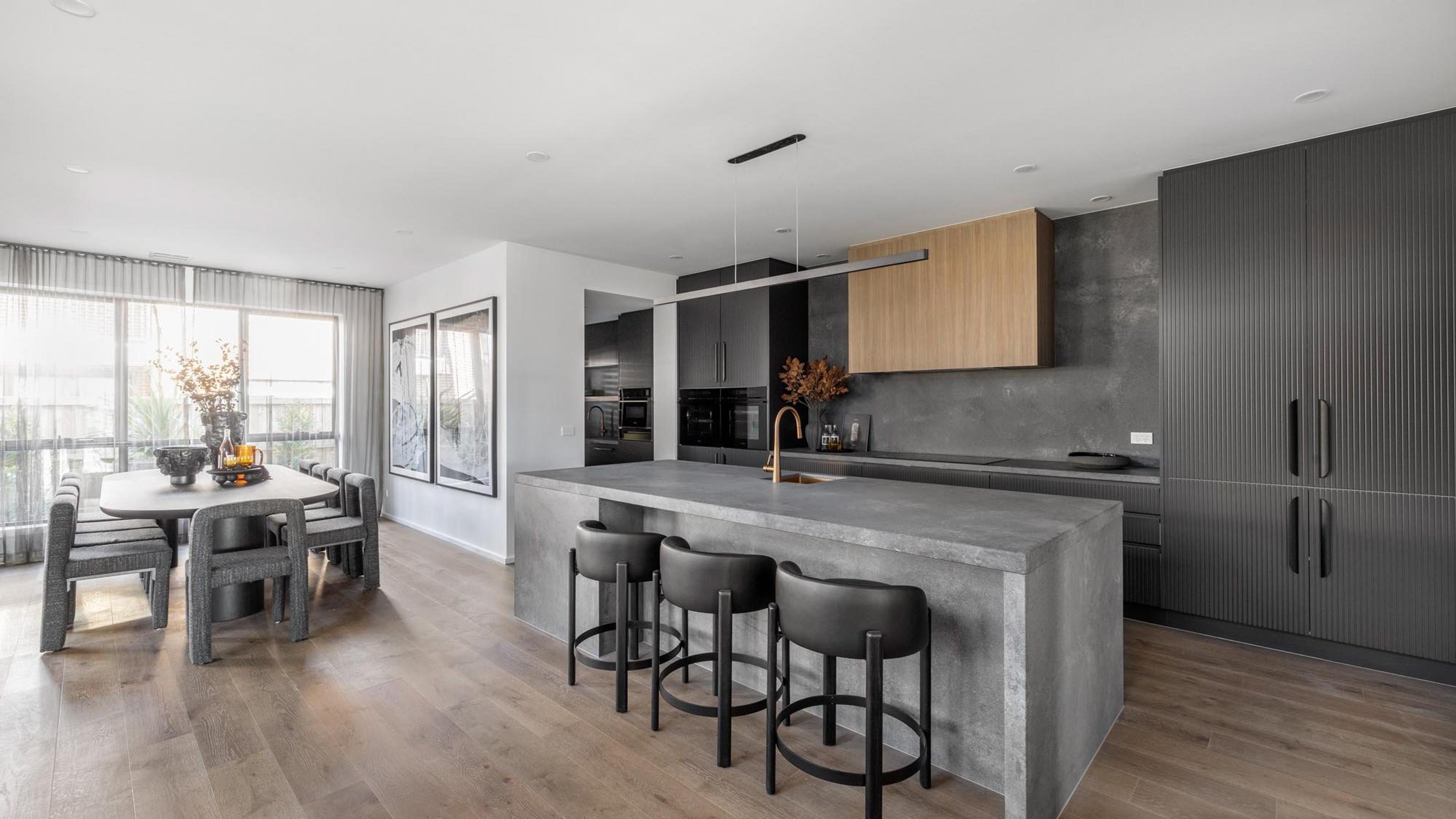An open-plan kitchen with a long stone island bench, bar stools and a dining table beyond