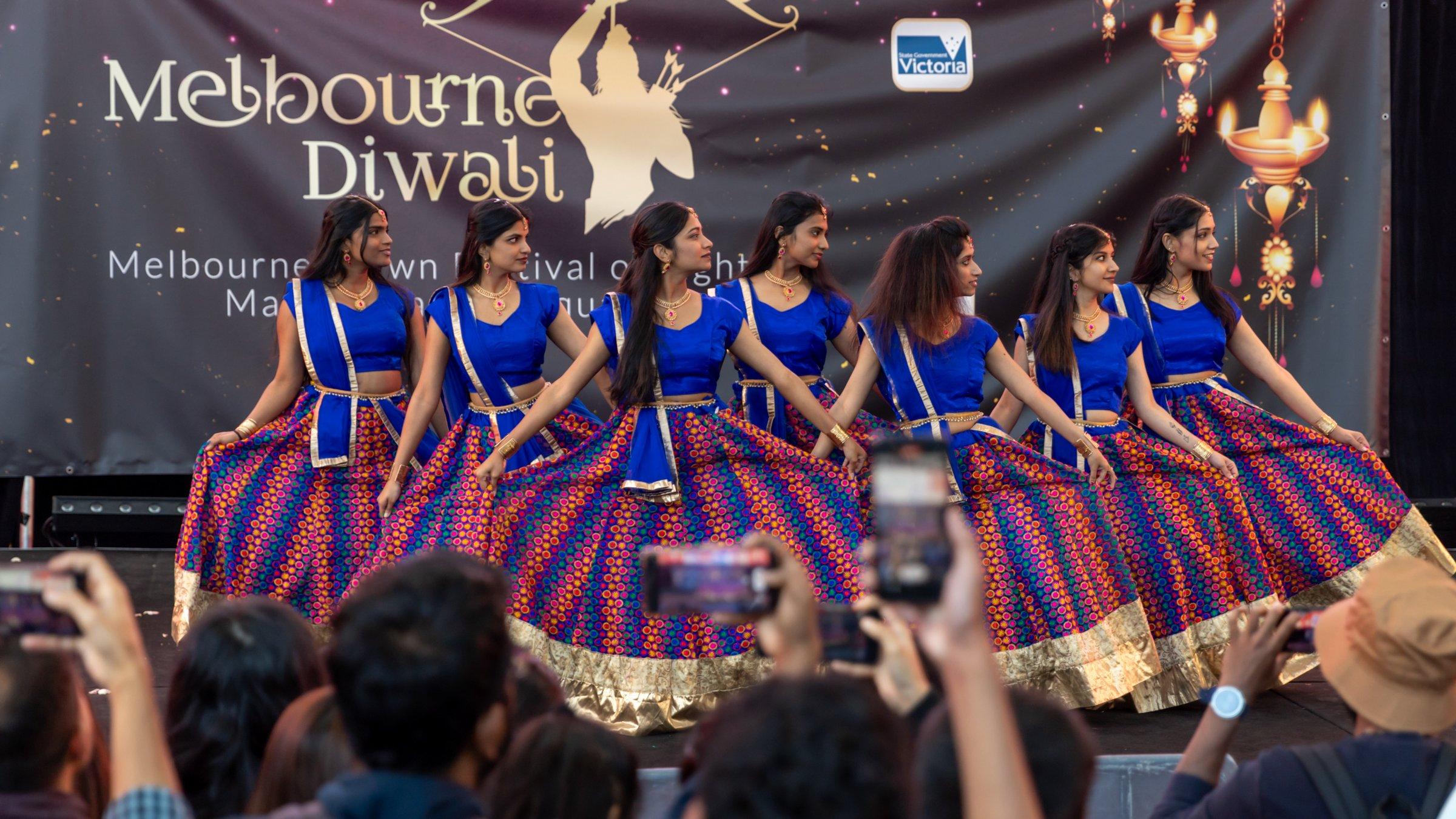 A dance group in blue and gold lehengas performing on the Melbourne Diwali stage as the audience films on their phones