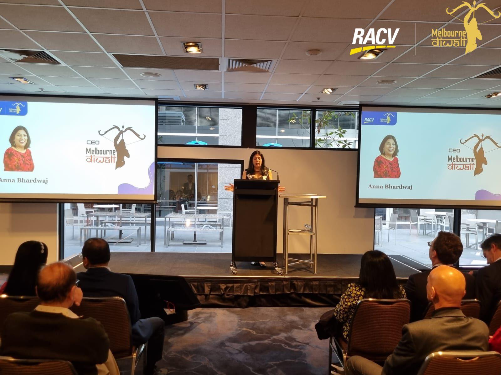 Anu Bhardwaj, CEO of Melbourne Diwali, at the lectern with her name slide on the twin screens