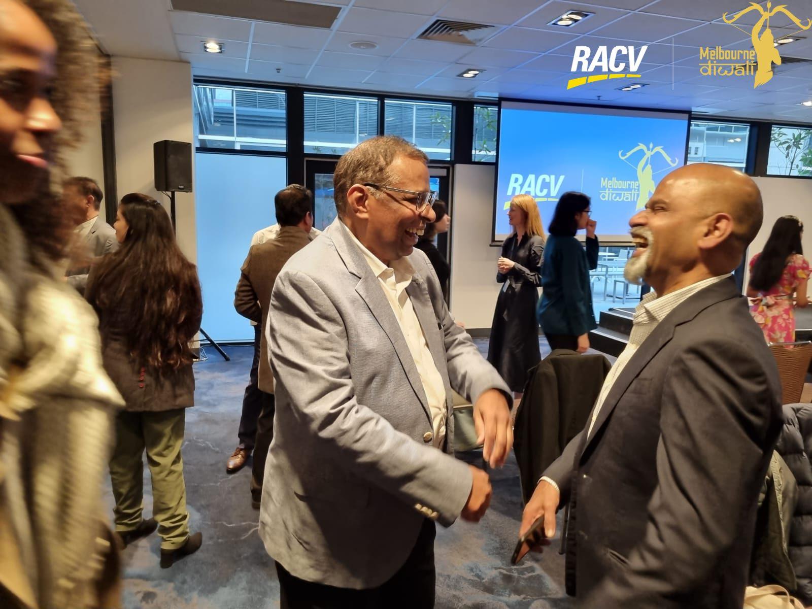 Two guests laugh as they greet each other in front of the RACV and Melbourne Diwali screen