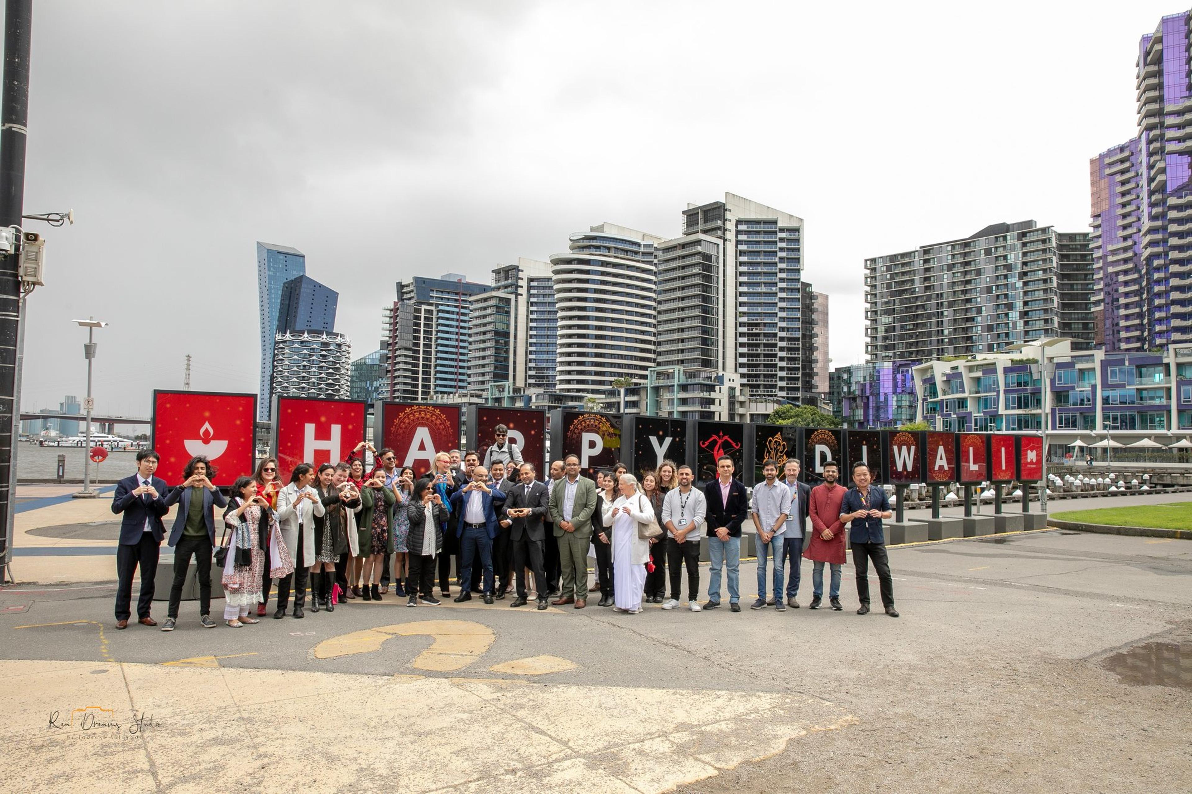 Guests at the HAPPY DIWALI sign on Harbour Esplanade, many making heart shapes with their hands, with Docklands towers behind