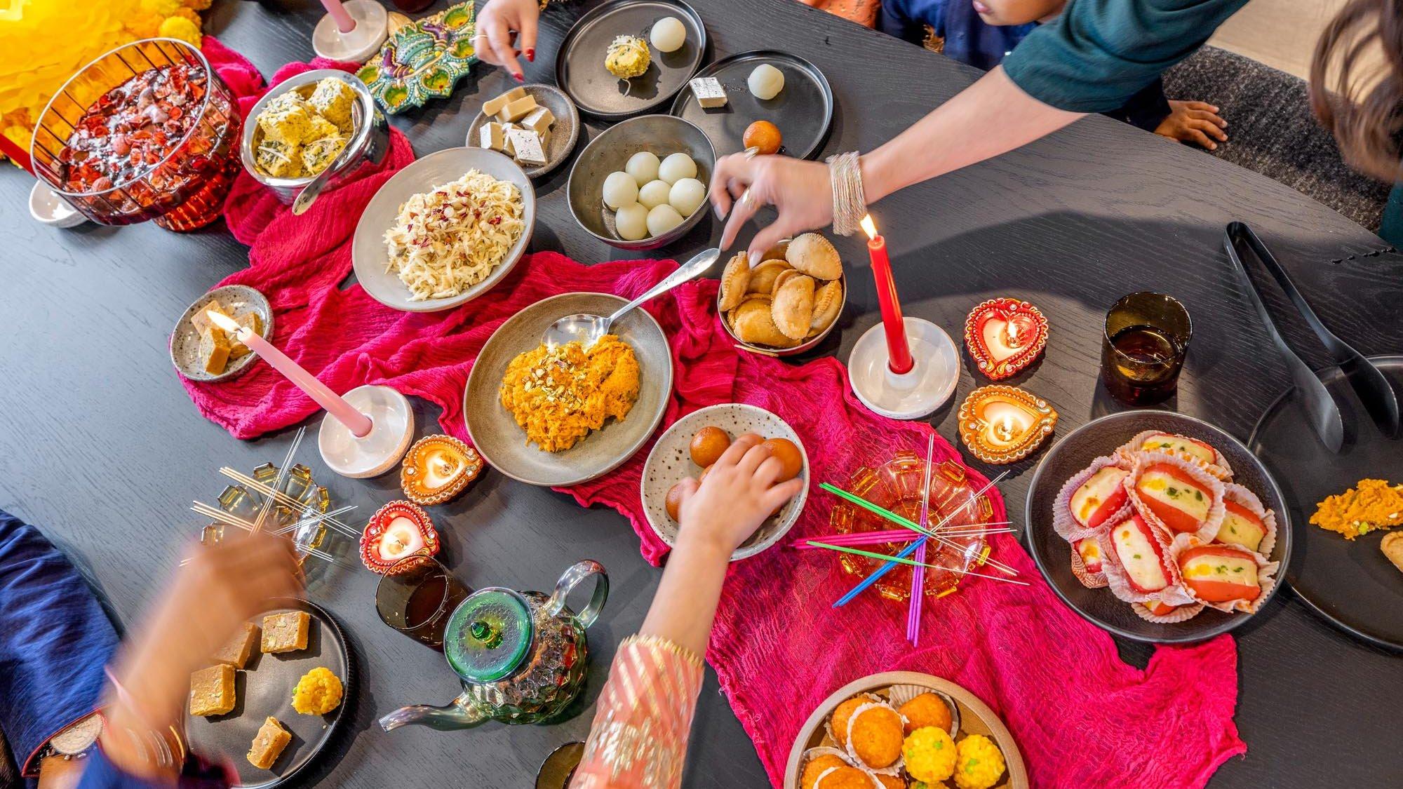 Overhead view of a Diwali table with sweets, diyas and family hands reaching in