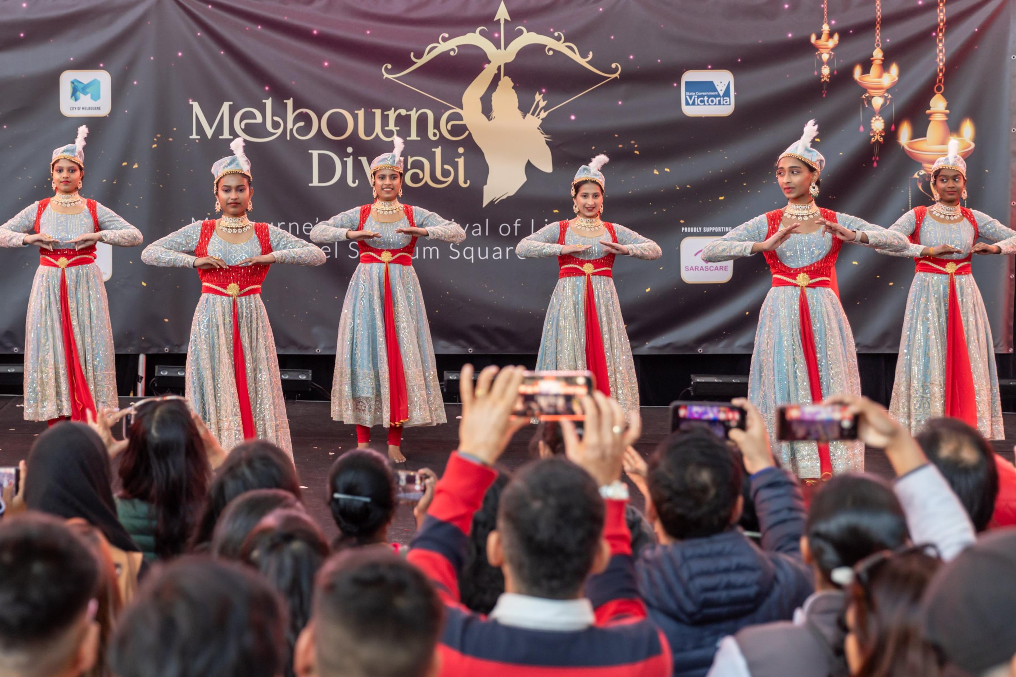 Dancers in silver and red costumes performing on the Melbourne Diwali stage at Marvel Stadium in 2023, with the audience filming on their phones