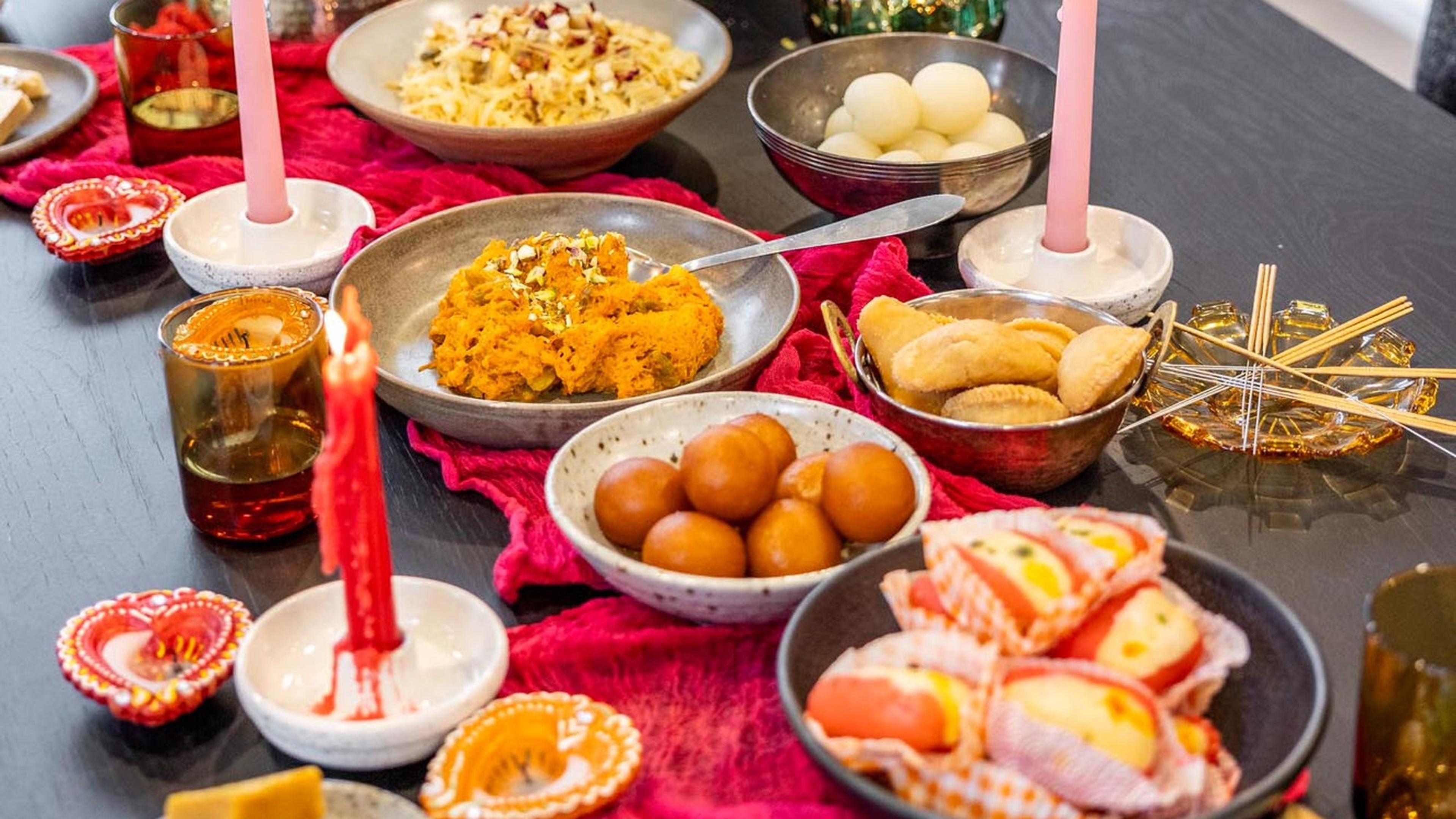 A Diwali table spread with sweets, diyas and red candles on a pink runner