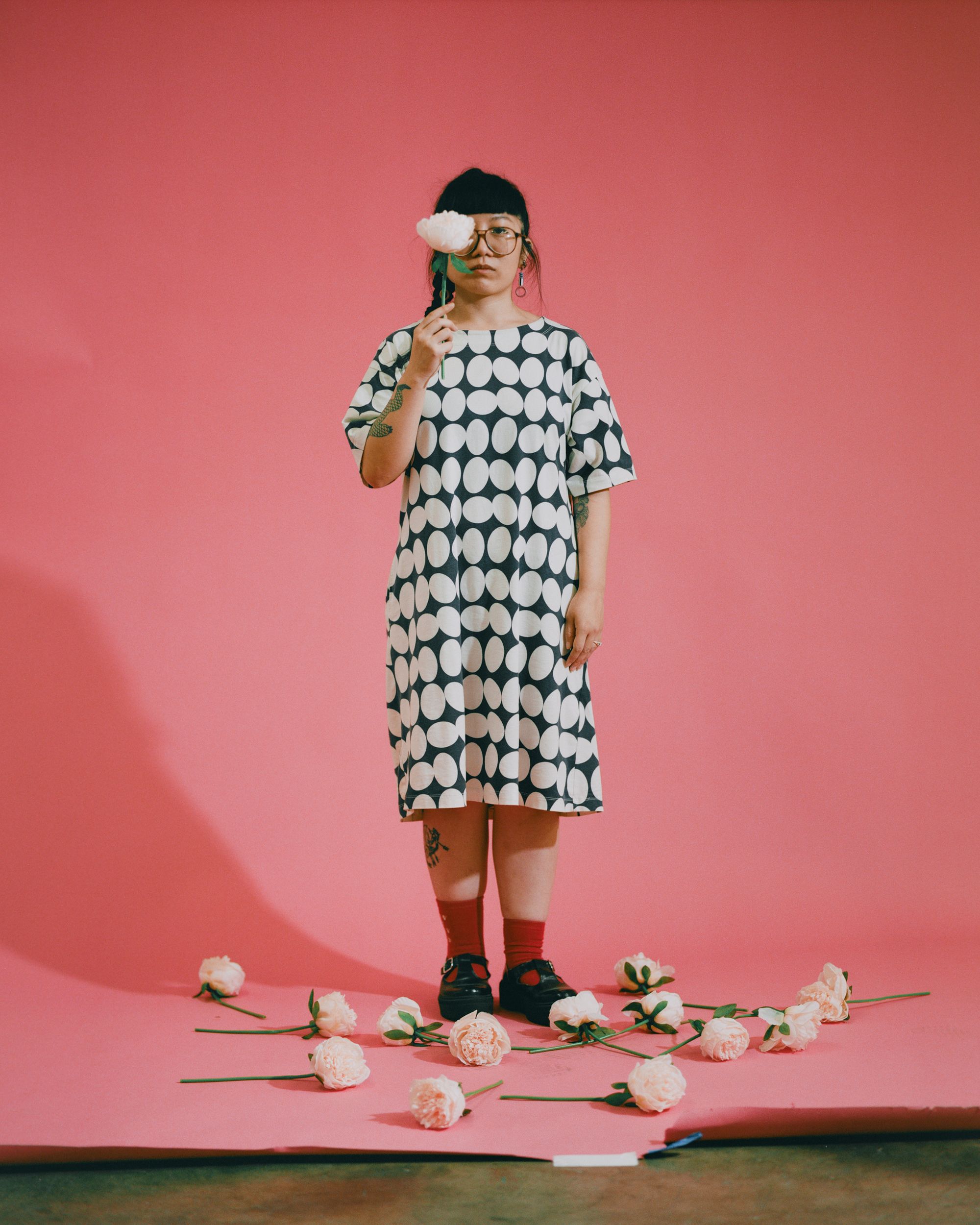 Photographic portrait of painter and illustrator Kristen Liu-Wong standing against a pink backdrop, surrounded by peonies.