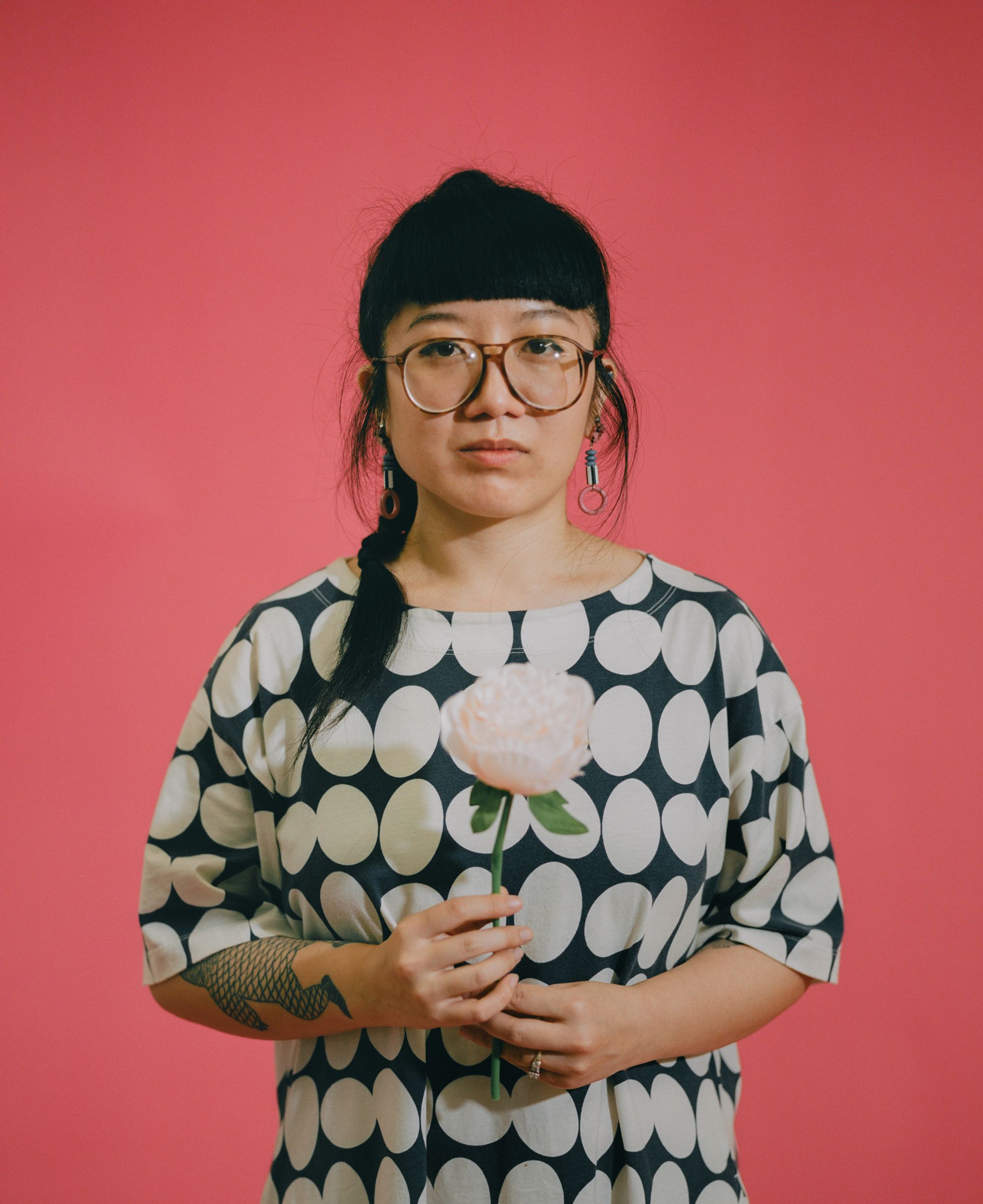 Photographic portrait of painter and illustrator Kristen Liu-Wong shot against a pink backdrop, holding a peony stem.