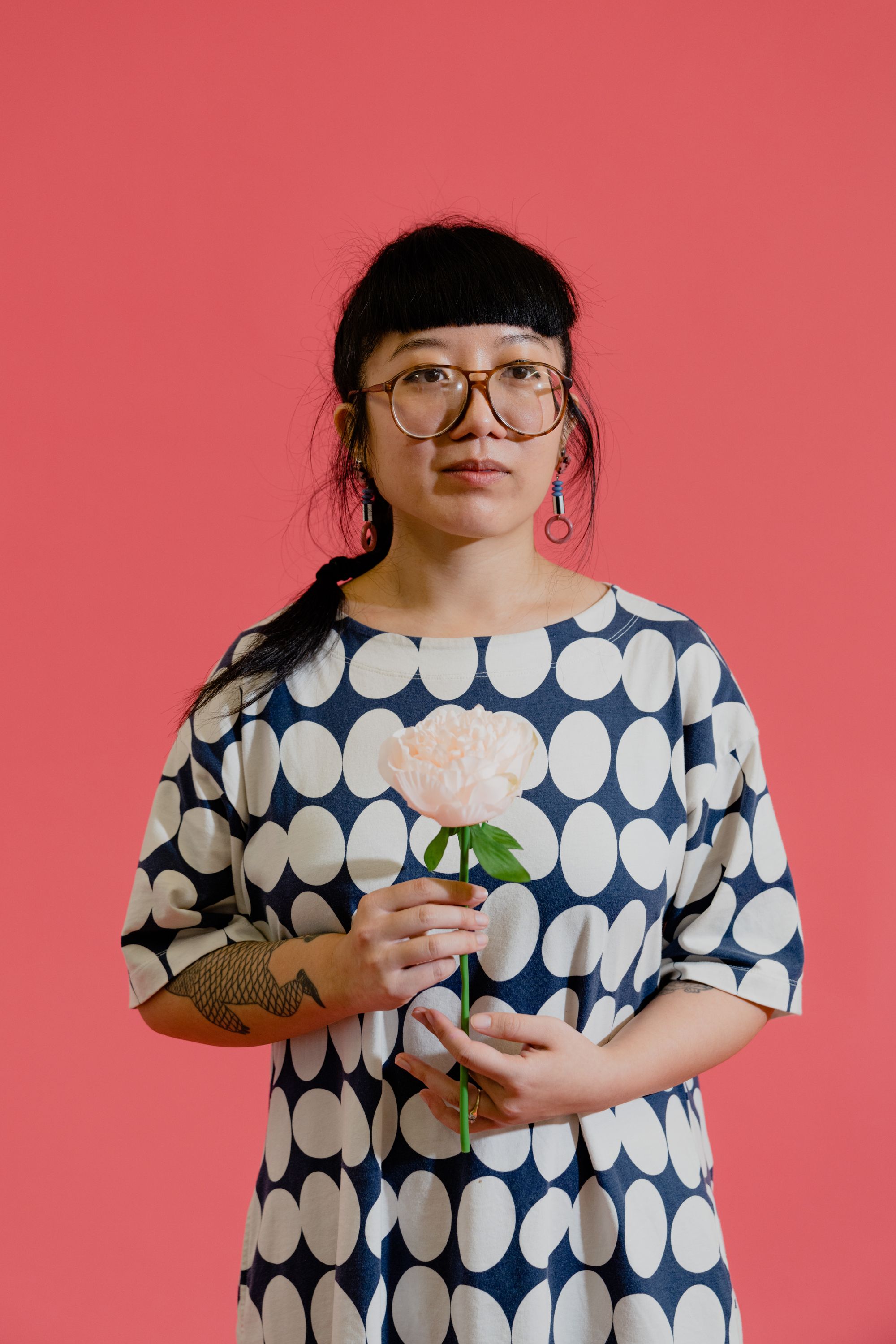 Photographic portrait of painter and illustrator Kristen Liu-Wong standing against a pink backdrop holding a peony stem.
