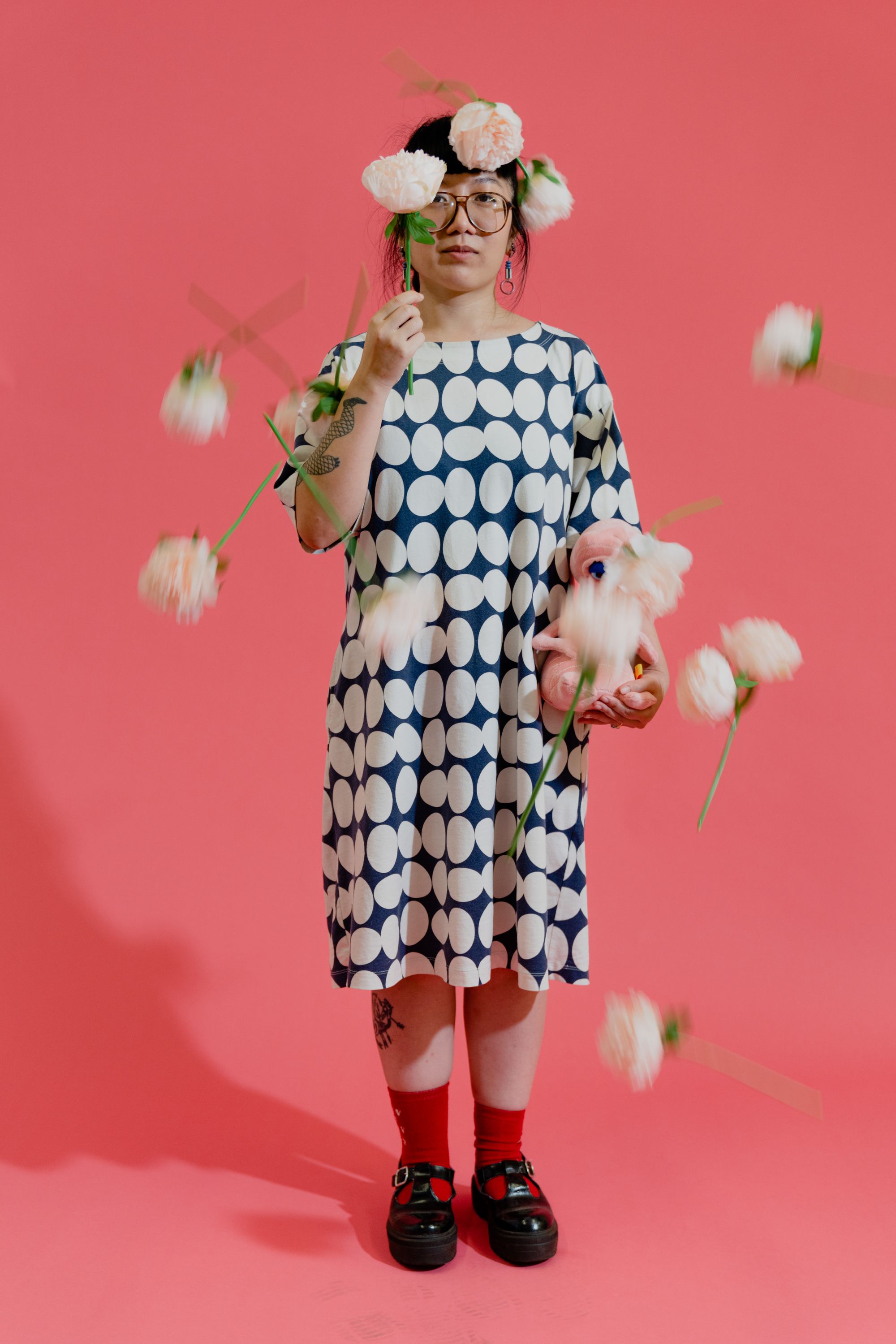 Photographic portrait of painter and illustrator Kristen Liu-Wong standing against a pink backdrop with falling peonies captured in action around her body.