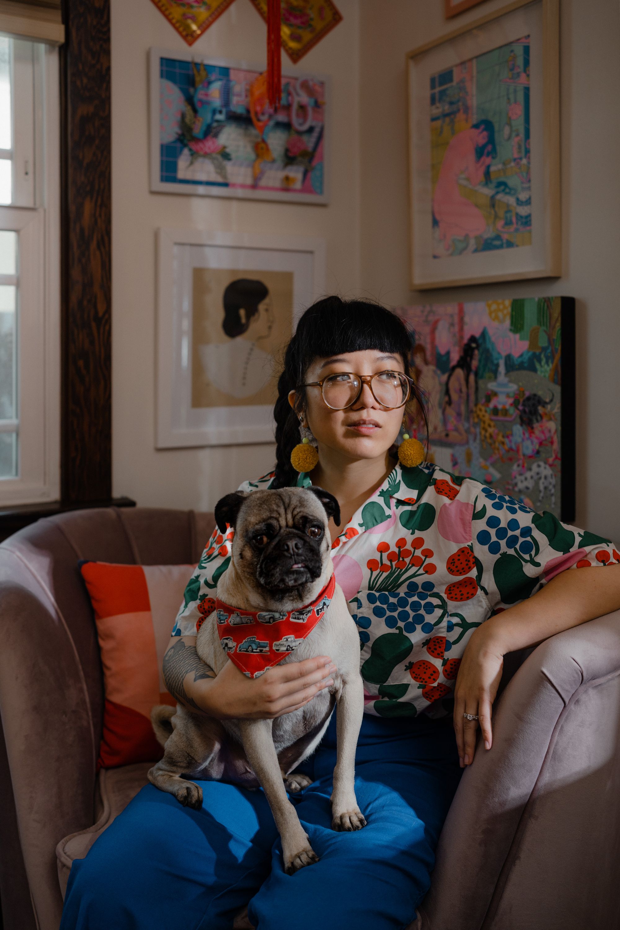 Photographic portrait of painter and illustrator Kristen Liu-Wong sitting on a plush chair in her home studio holding her pet pug named Rooster.