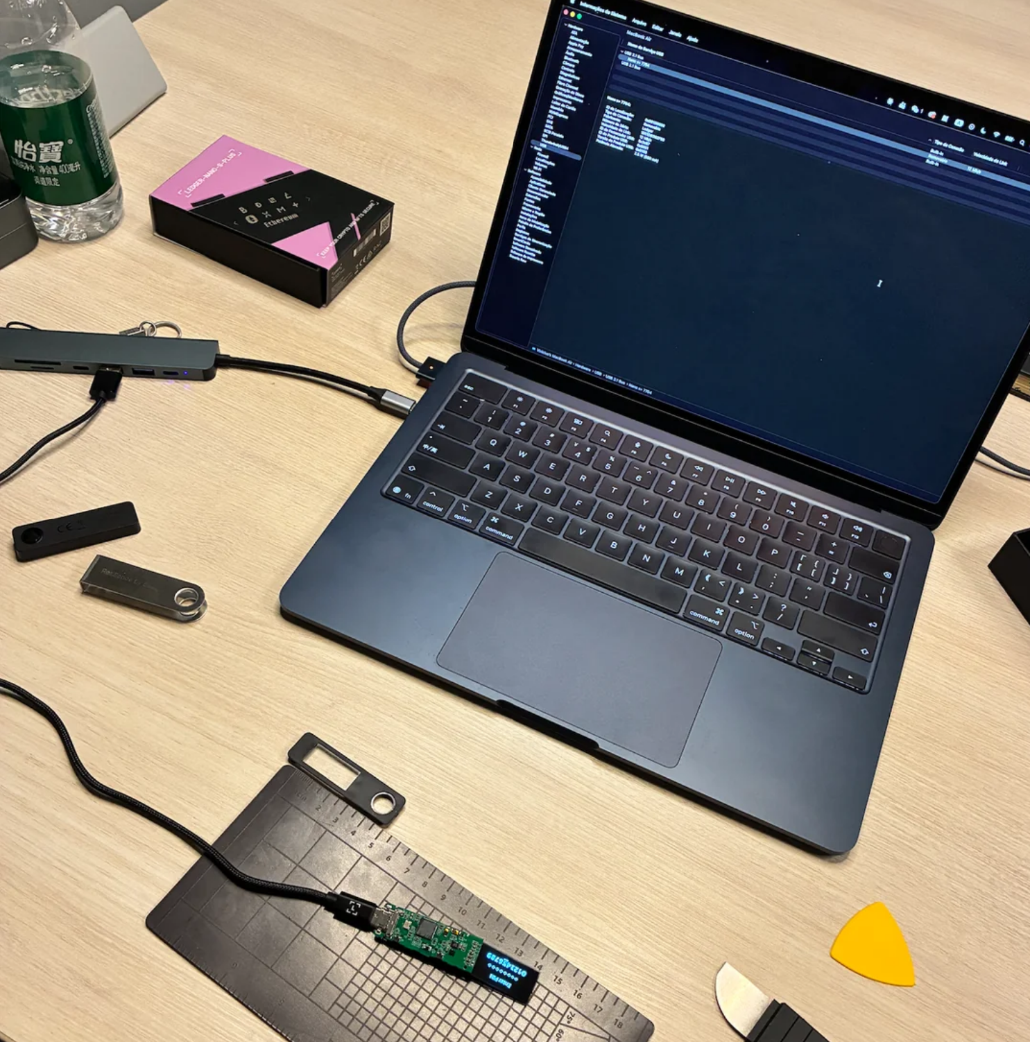 A MacBook on a wooden desk surrounded by hardware analysis tools. The counterfeit device's exposed circuit board is connected via USB cable to a workstation mat in the foreground, with analysis software open on the laptop screen. The fake Ledger's pink and black retail packaging is visible in the top left corner, alongside what appears to be a genuine Ledger device.