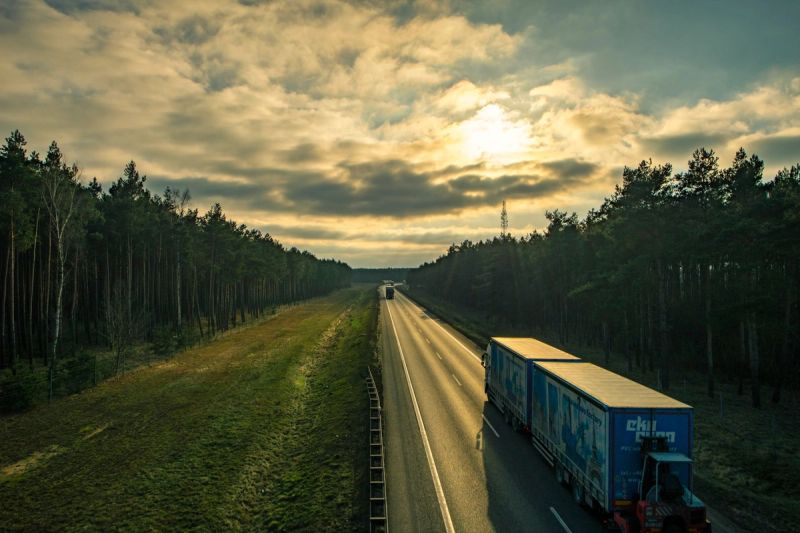 Une route bordée de forêt au coucher du soleil, avec des camions au loin.