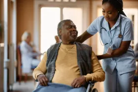 Older Black man sits in wheelchair, smiling up at female Black nurse as she reaches toward him, also smiling