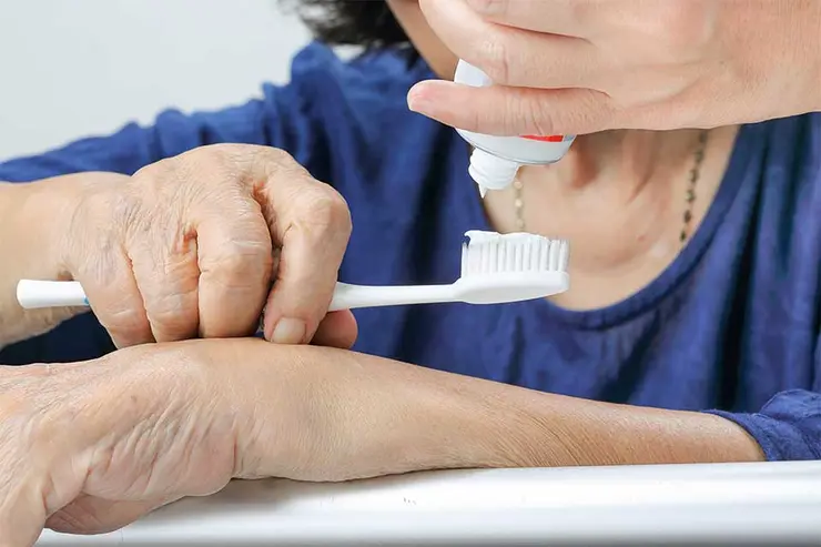 Elderly woman receiving help putting toothpaste on her toothbrush from a caregiver.