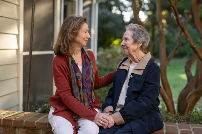 An older woman and a younger woman sitting together outdoors, holding hands and smiling at one another.