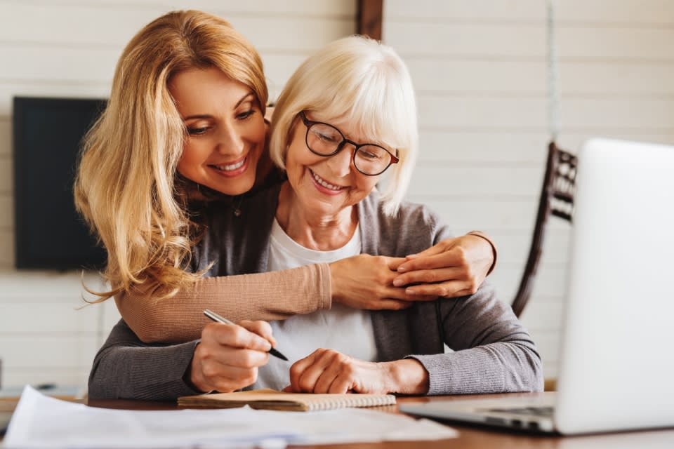 Middle-aged woman hugs her mother from behind. Elder mother sits in front of a laptop with documents in front of her and wearing glasses.