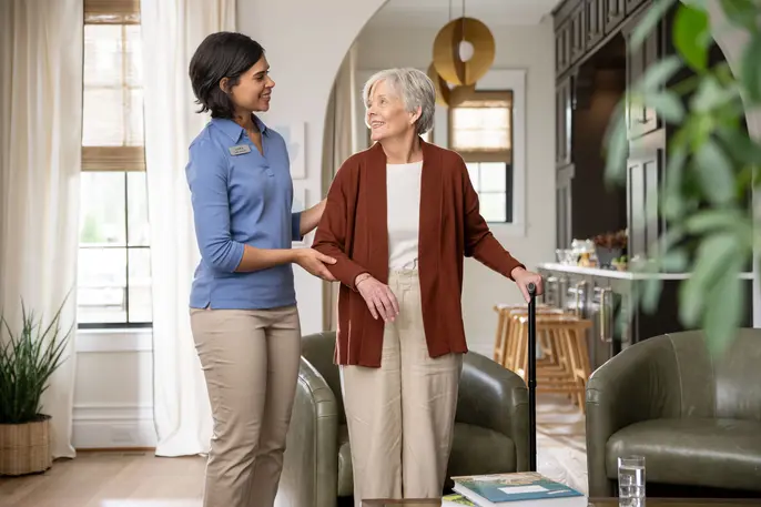 Caregiver supports an older woman walking with a cane in an assisted living community.