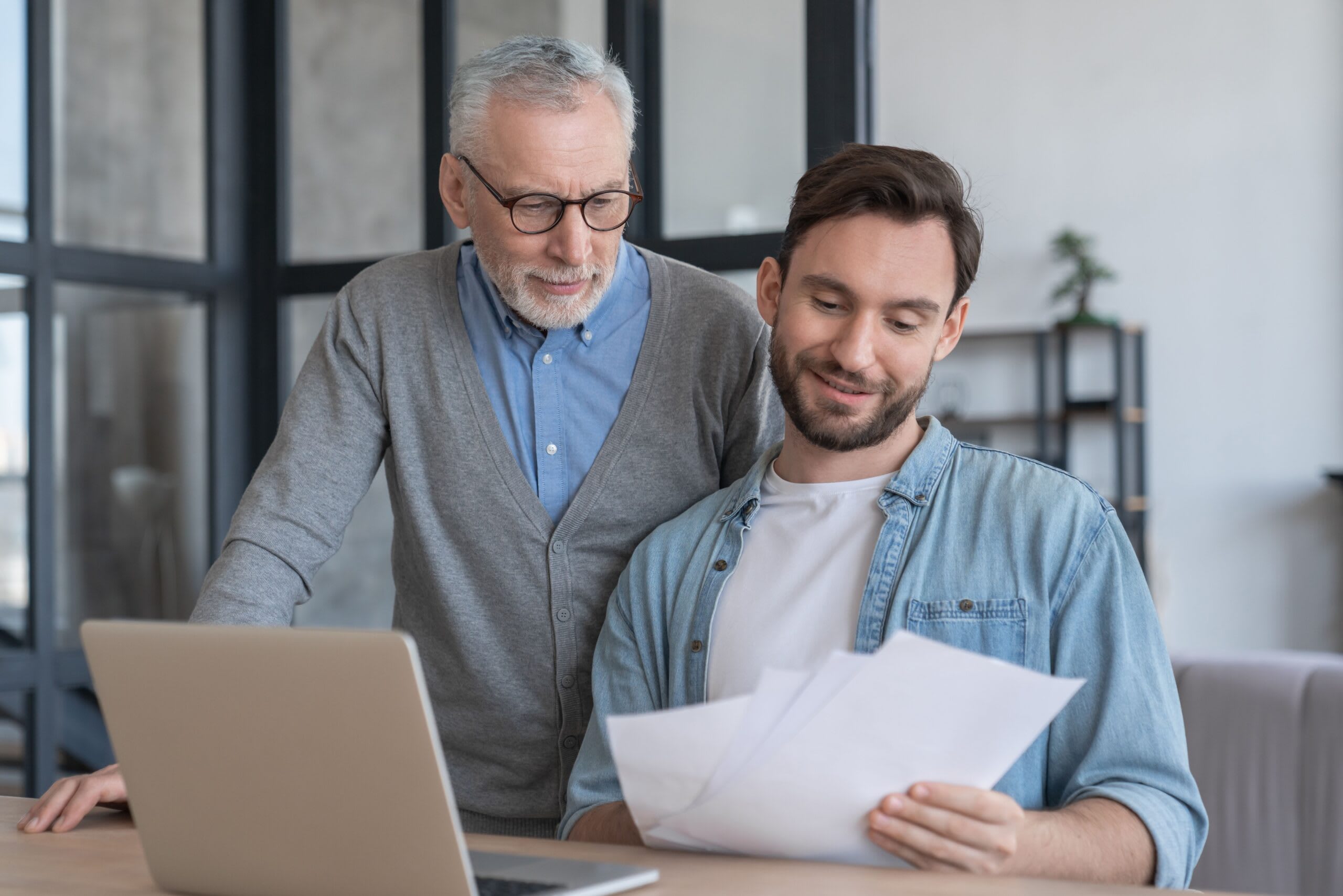 Adult son smiles as he helps his senior father paying for home care at home in front of a laptop on a table