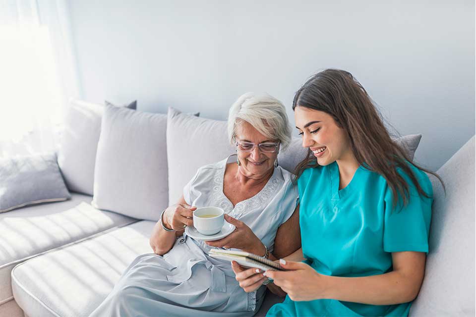 Elderly woman being read to by her private caregiver while sitting on a couch.