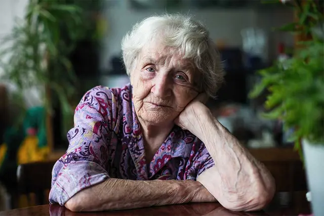 Elderly woman seated at a table resting her head on her hand.