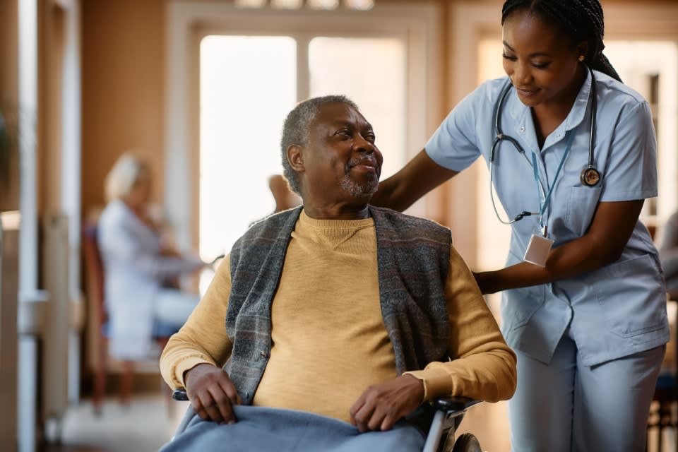 Older Black man sits in wheelchair, smiling up at female Black nurse as she reaches toward him, also smiling