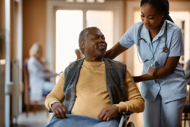 Older Black man sits in wheelchair, smiling up at female Black nurse as she reaches toward him, also smiling