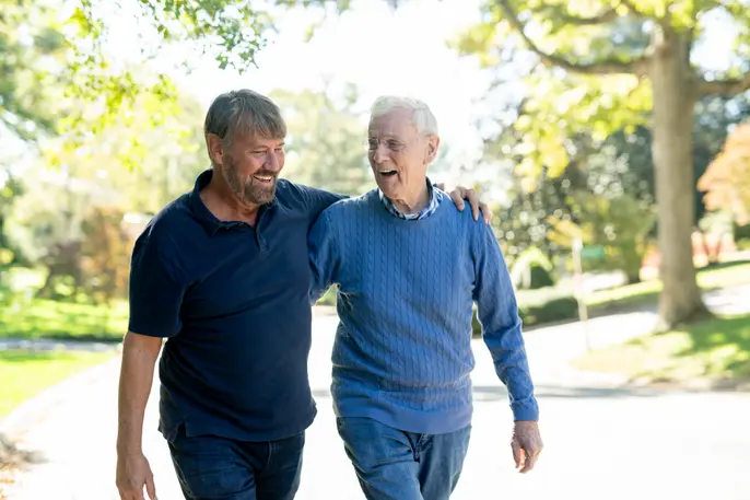 A photo of an older man and another man walking arm in arm on a sunny, tree-lined path, smiling and talking outdoors.