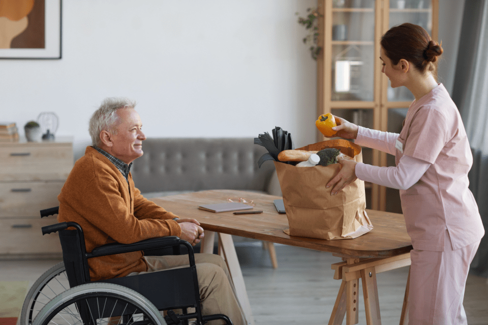 A senior man sits at his table and smiles while his in-home caregiver brings his groceries.