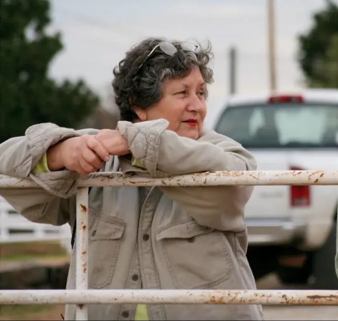Martha leans on a fence at an equestrian venue, looking toward the horse arena.