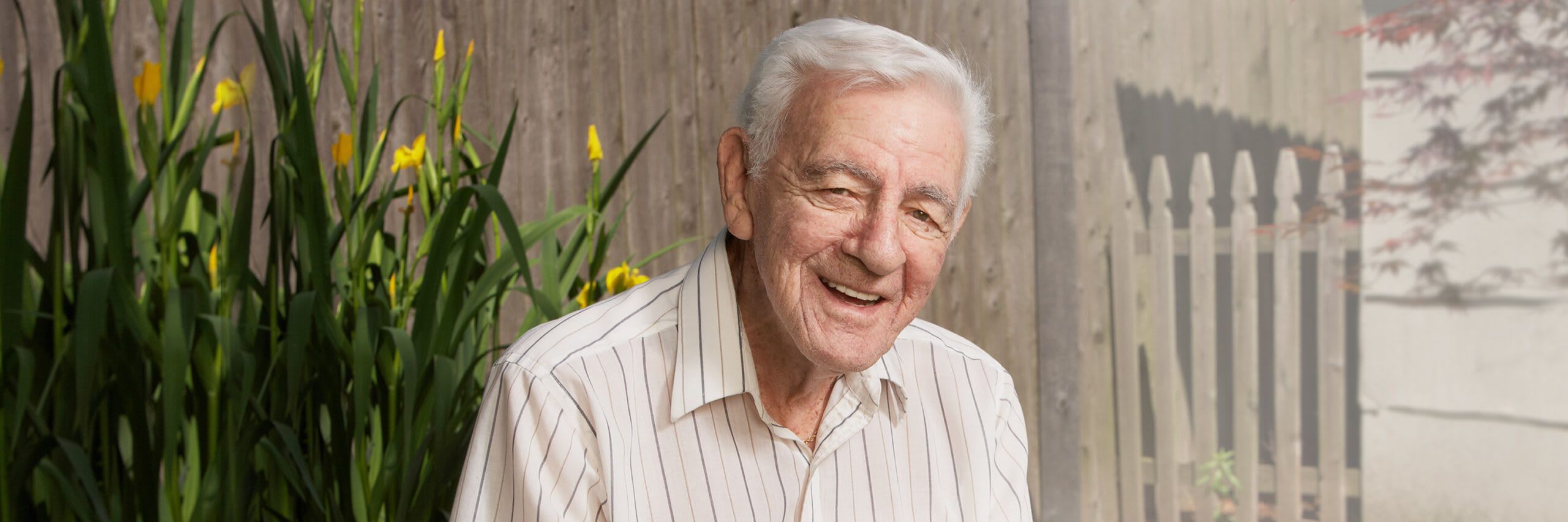 A senior man smiling in front of flowers outside his house
