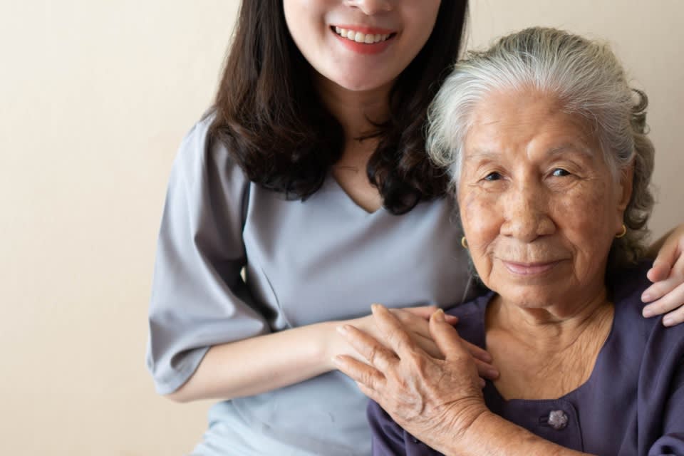 Older Asian woman smiles gently into the camera while holding the hand of a young Asian woman, also smiling