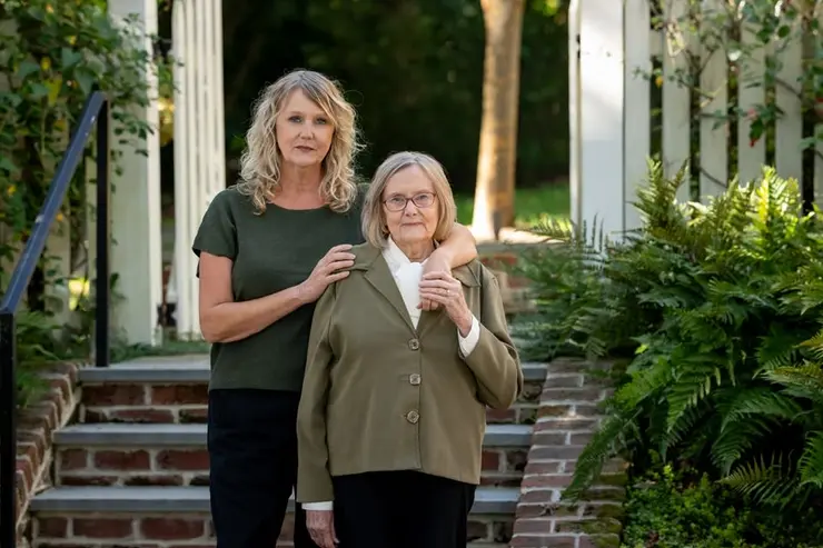 Adult daughter stands with her older mother outside a home, representing family caregiving and aging at home.