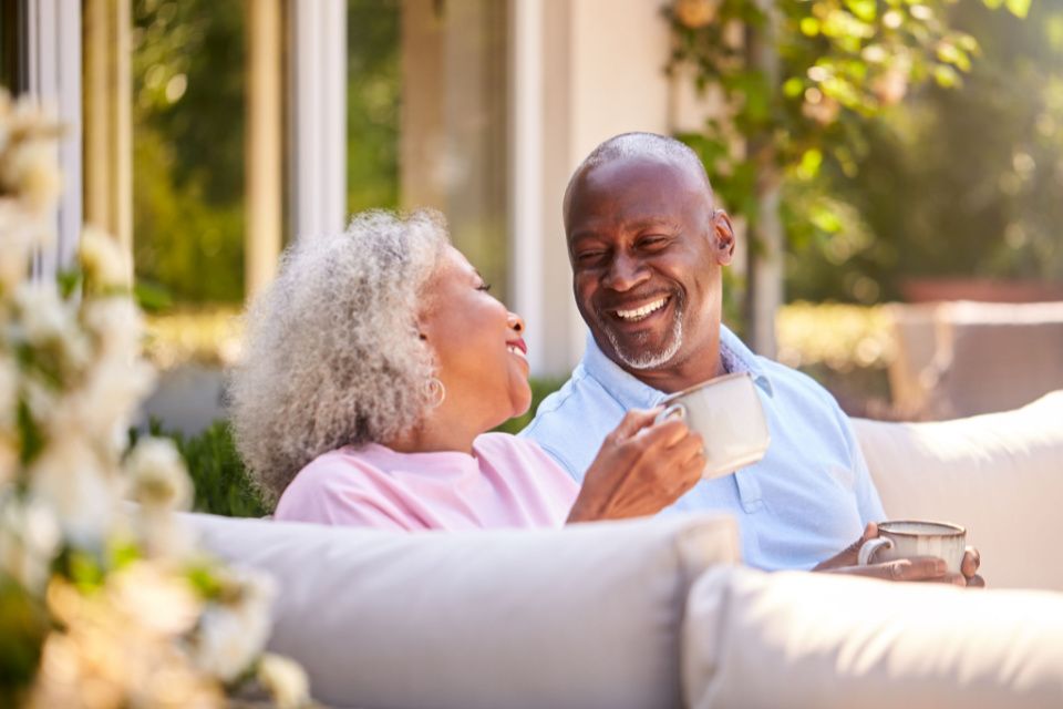 A senior couple sitting on a porch, holding mugs and smiling at each other
