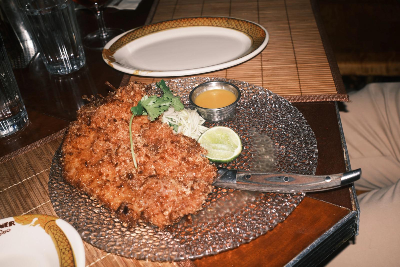 A large craggy breaded chicken schnitzel on a hammered silver charger with a sprig of coriander, a small pile of shredded papaya salad, a lime wedge, and a metal pot of orange dipping sauce, a steak knife laid across it and an empty snake-rimmed oval plate on the bamboo mat behind.