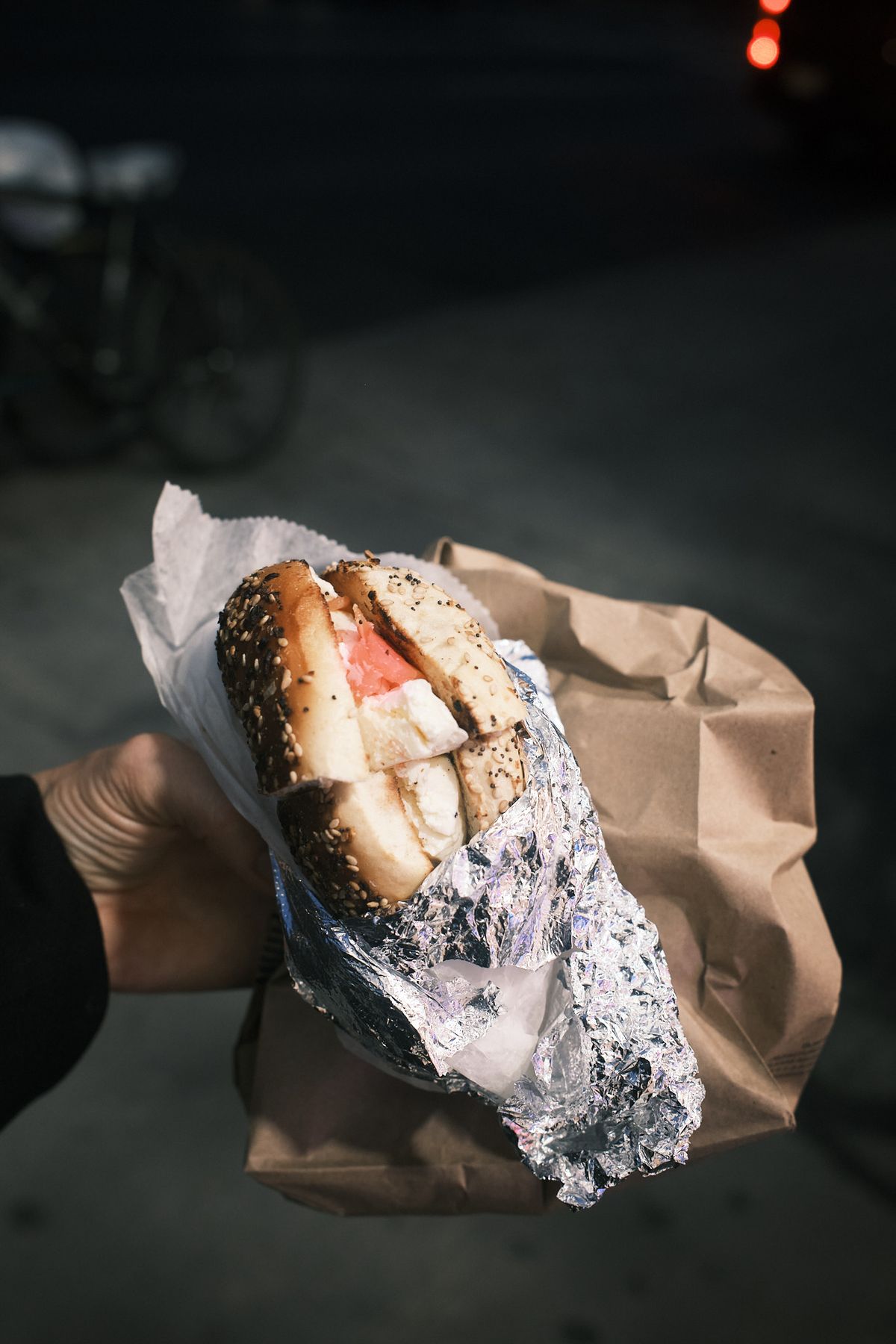 A hand holding a seeded everything-bagel filled with smoked salmon and thick cream cheese, the foil and paper peeled back over a brown paper bag, on a dark street at night with a parked bicycle and a red tail light blurred behind.