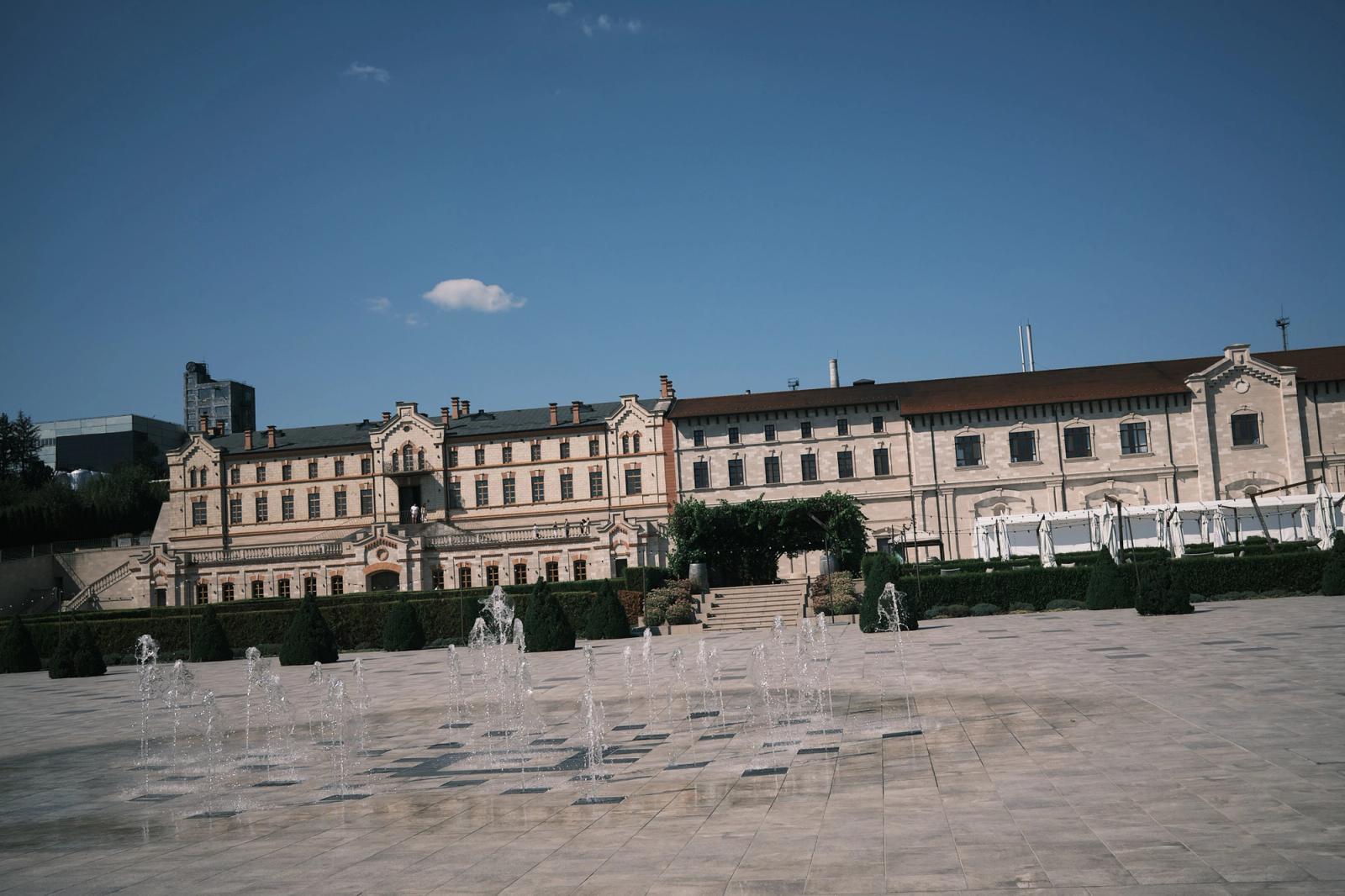 The long pale limestone facade of Castel Mimi under a deep blue sky, with low water jets playing across the stone forecourt.