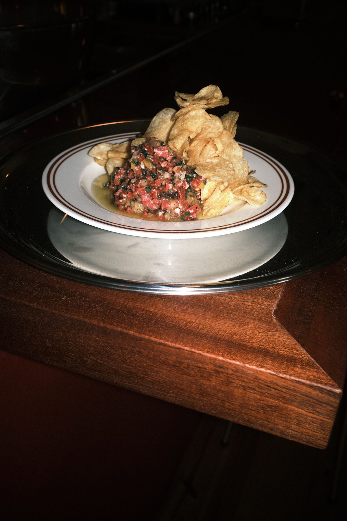 A mound of chopped beef tartare flecked with herbs and piled with salted crisps, on a brown-striped plate set on a silver charger at the edge of a dark polished wooden bar ledge.