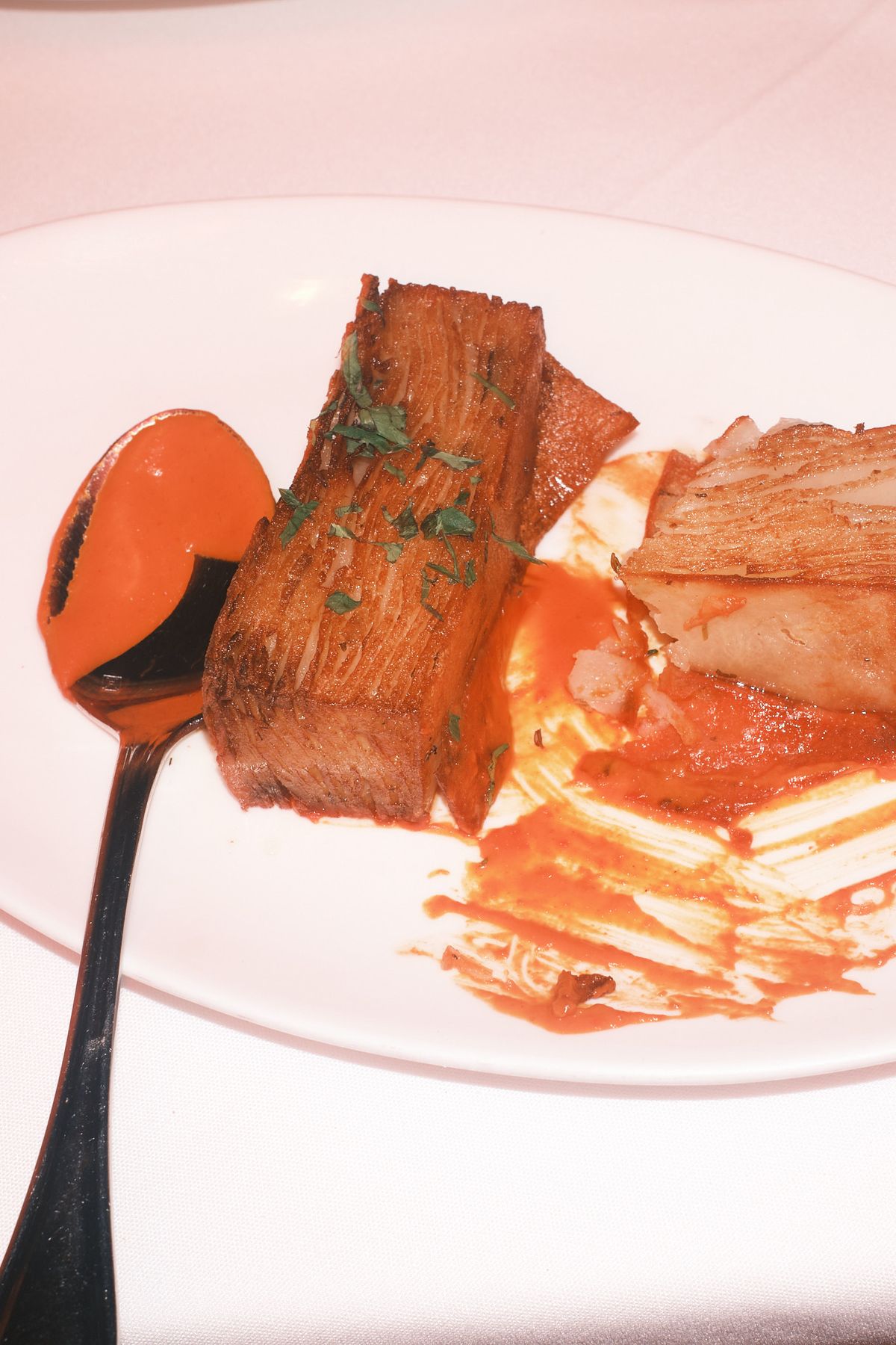 A rectangular block of finely layered potato, crisp and bronzed at the edges and scattered with chopped herbs, lying on a white plate beside a spoon of red romesco, with a second paler layered piece and a smear of sauce behind.