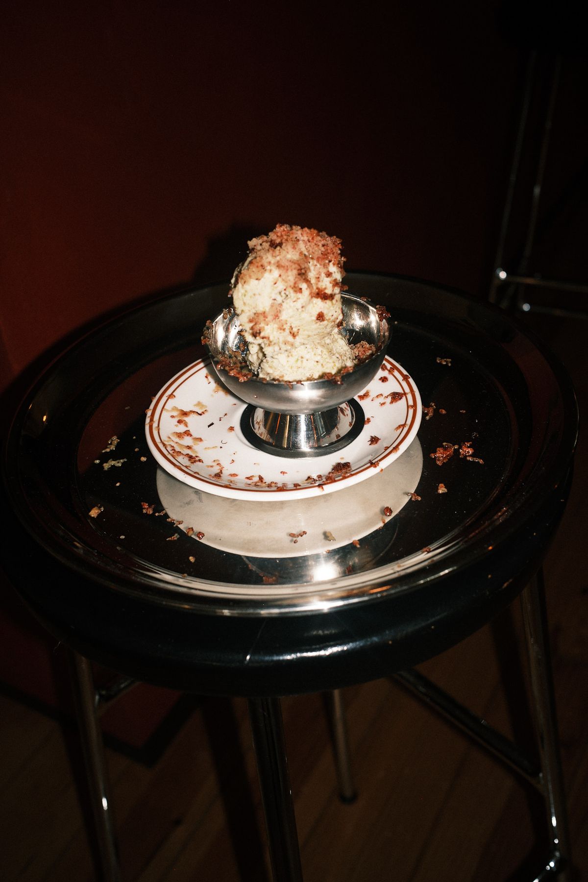 A scoop of pale ice cream heaped with rye crumb in a silver footed coupe, on a brown-rimmed saucer on a round black bar stool, crumbs scattered across the surface in low red light.